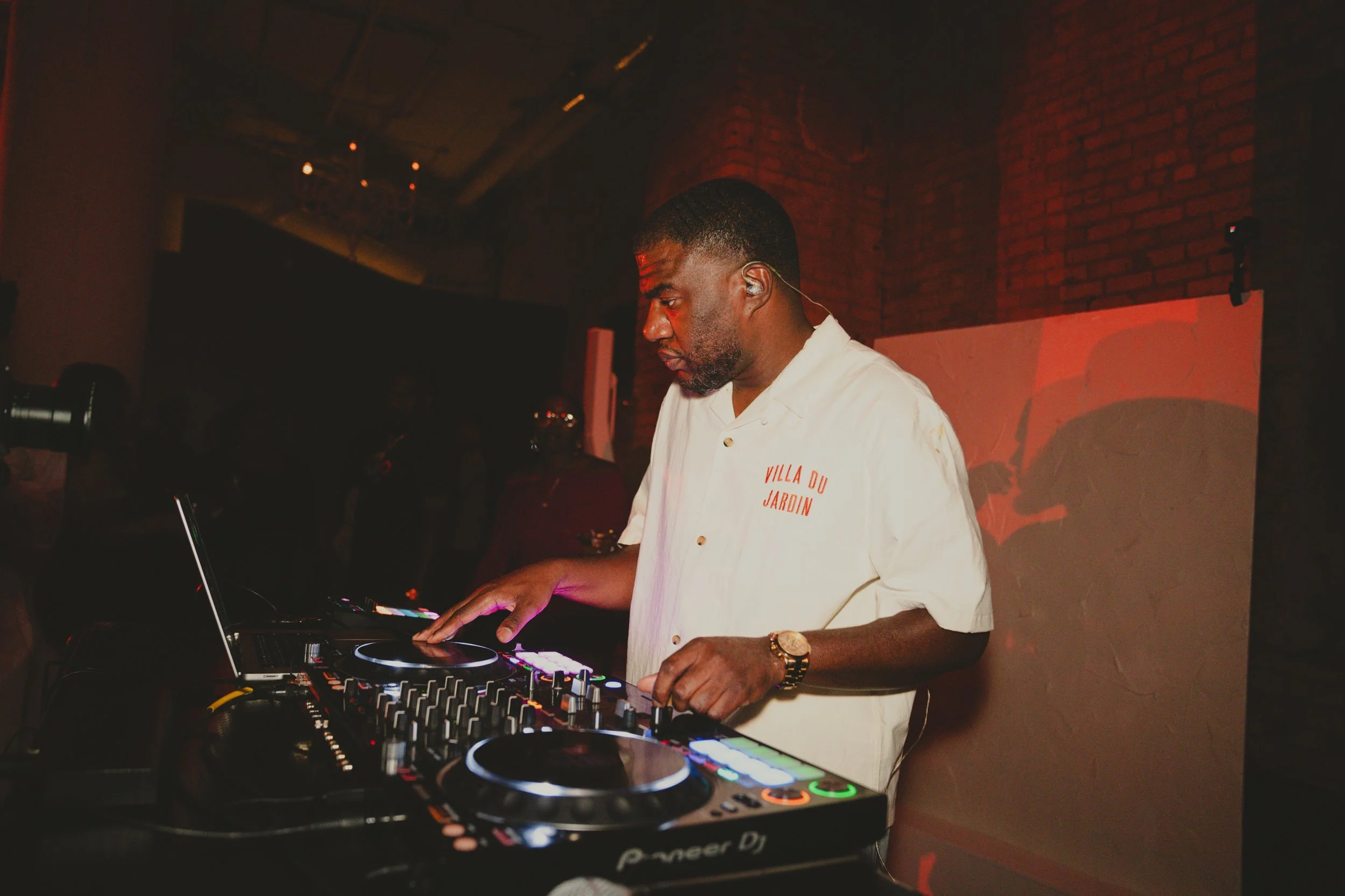 A DJ wearing a white shirt with red text on it, standing at a DJ turntable, in a dimly lit indoor space with a brick wall background.