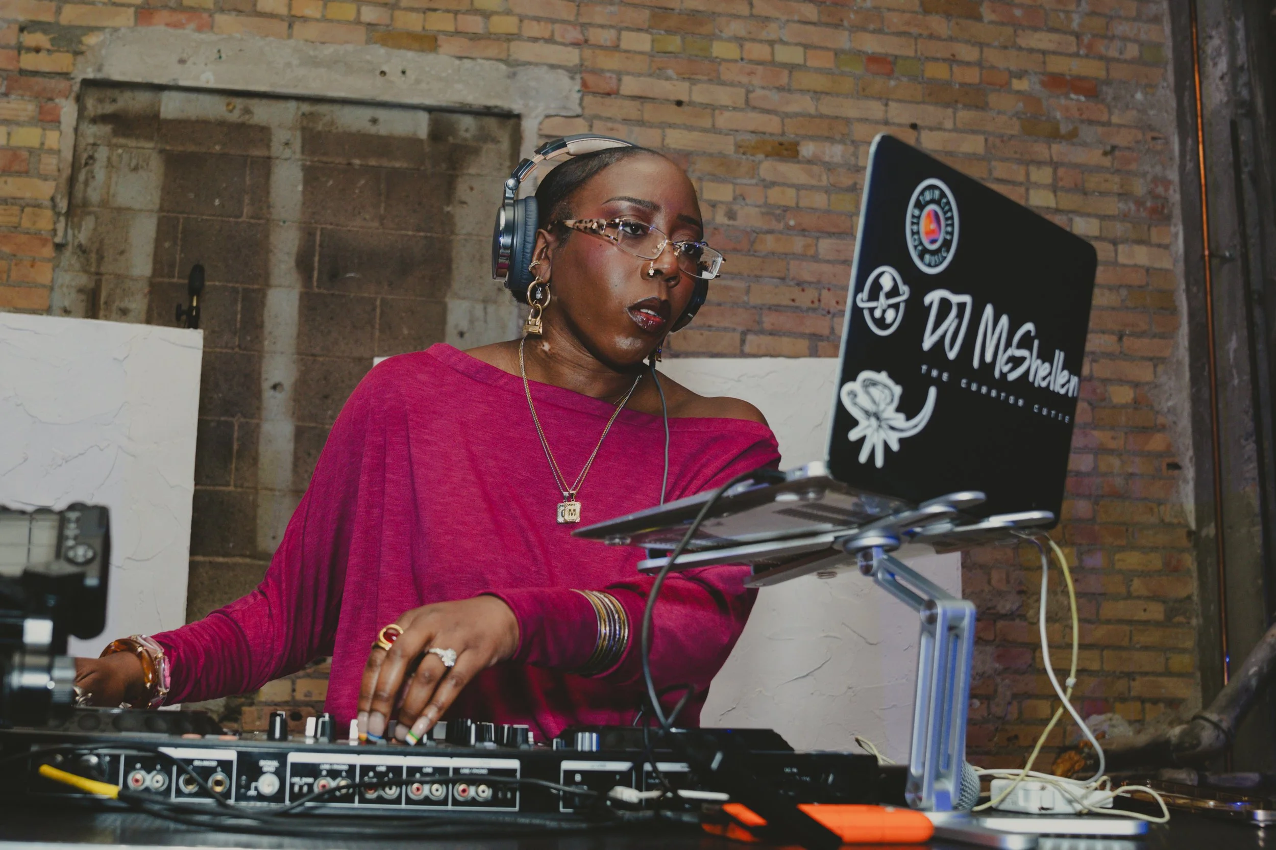 A woman in a pink top with glasses and jewelry, wearing headphones, is DJing with a mixer and laptop against a brick wall.