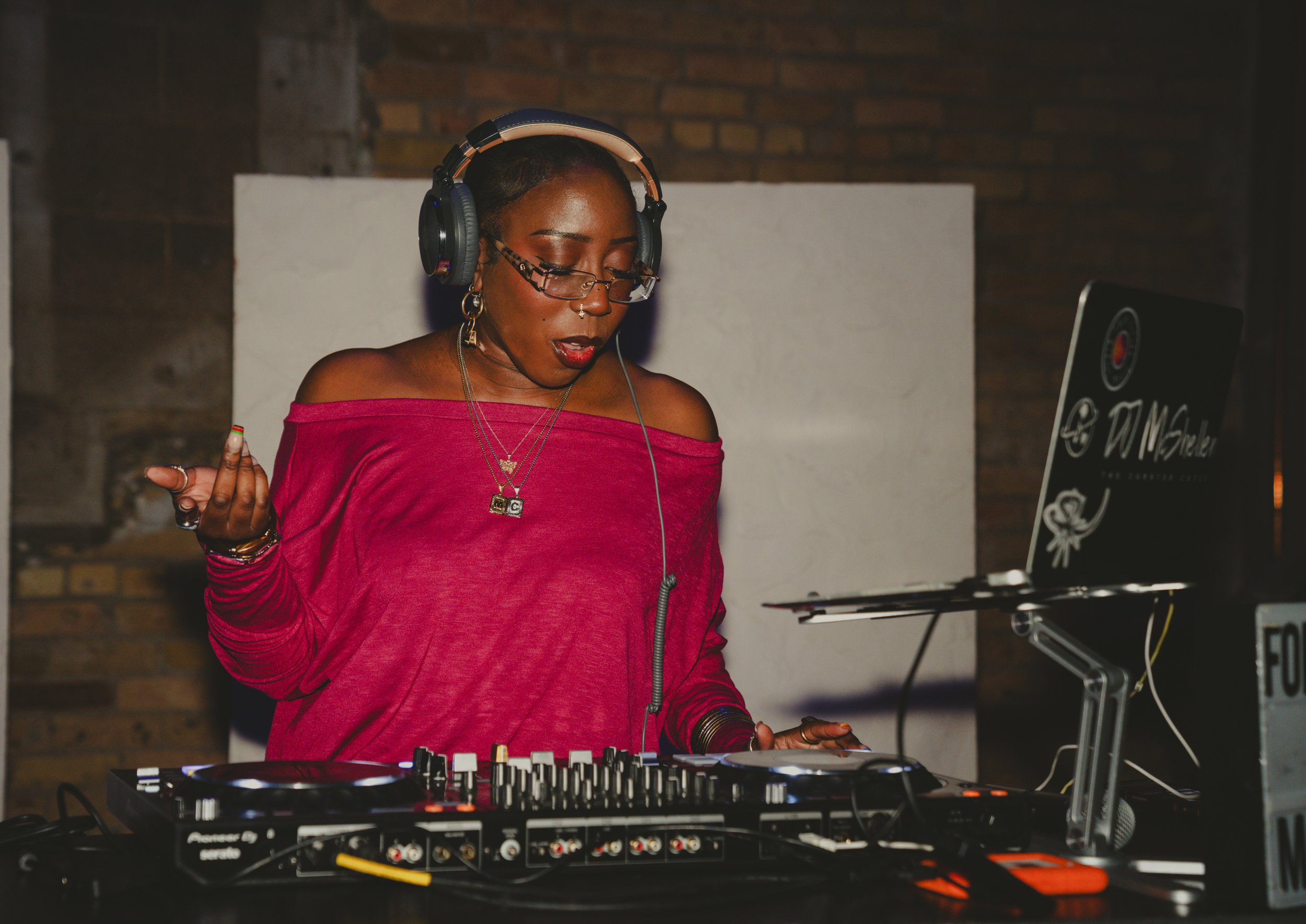 A woman with short hair, glasses, and earrings, wearing a pink off-shoulder top, is DJing with a turntable and mixer in front of her. She has headphones over her ears and appears to be enjoying the music.
