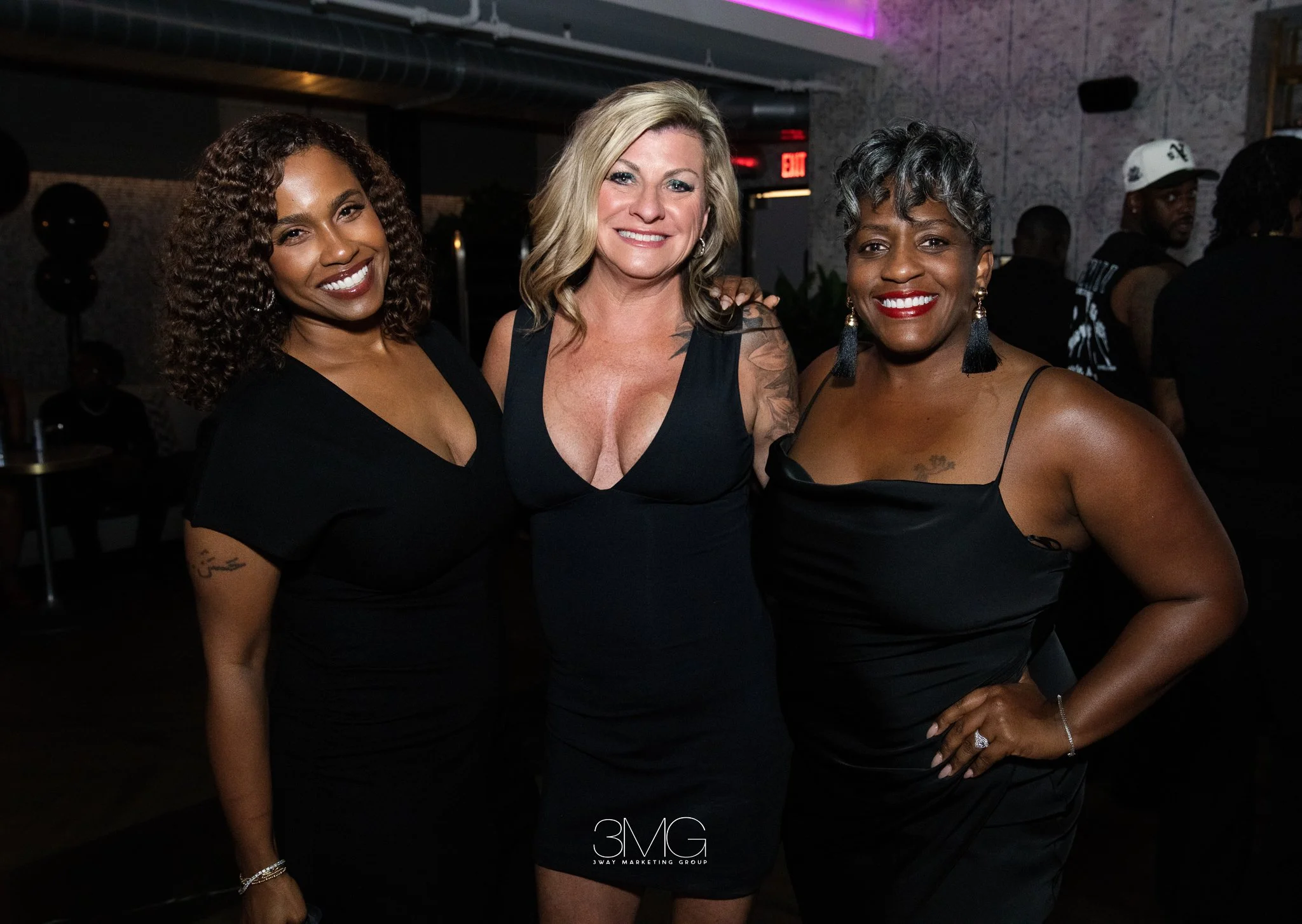 Three women smiling and posing together in a nightclub or party setting, dressed in black outfits, with the logo '3MG' at the bottom of the image.