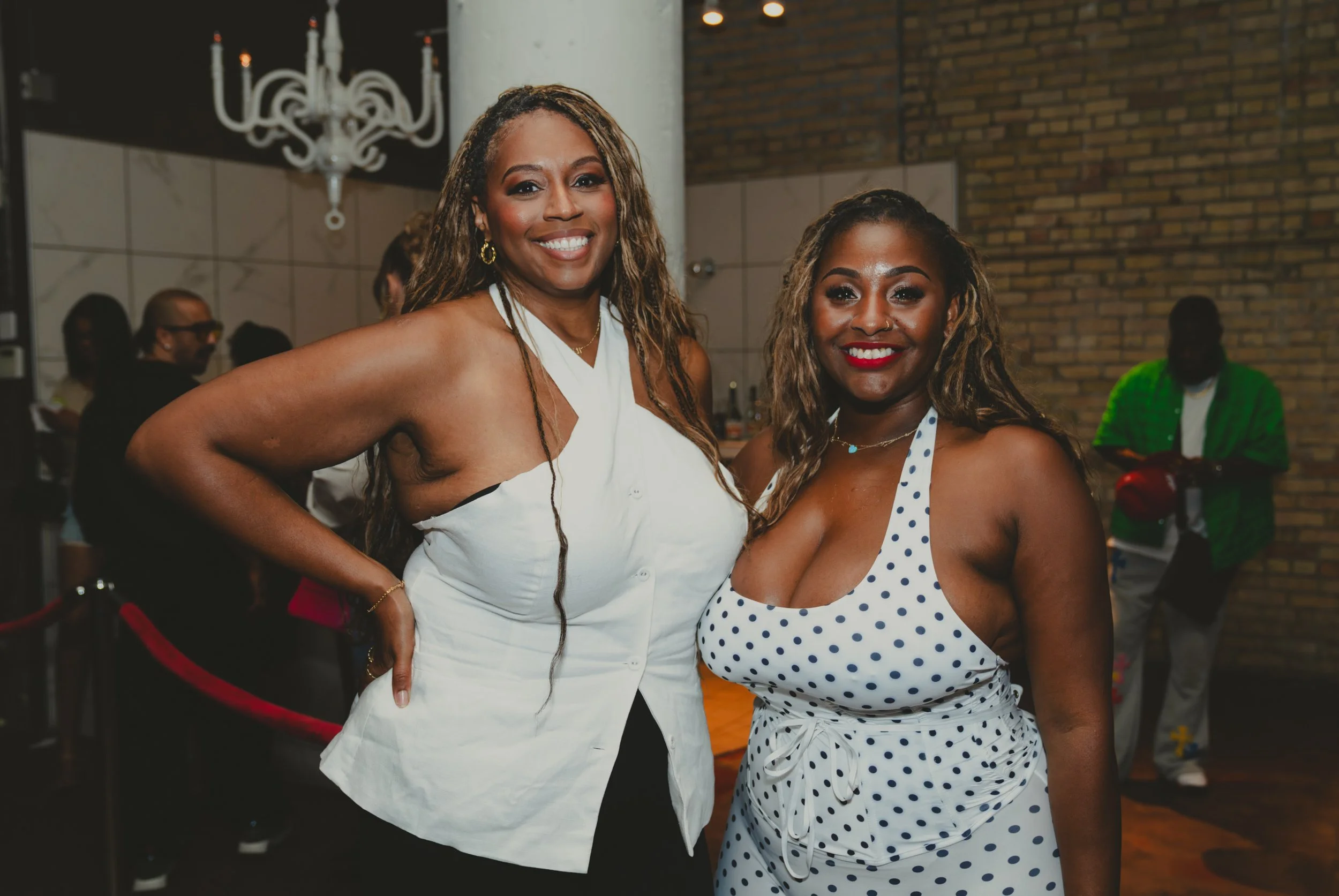Two smiling women posing together at an indoor social event, one wearing a white dress with ruffles and the other in a white polka-dot dress. Background shows other attendees and a brick wall.