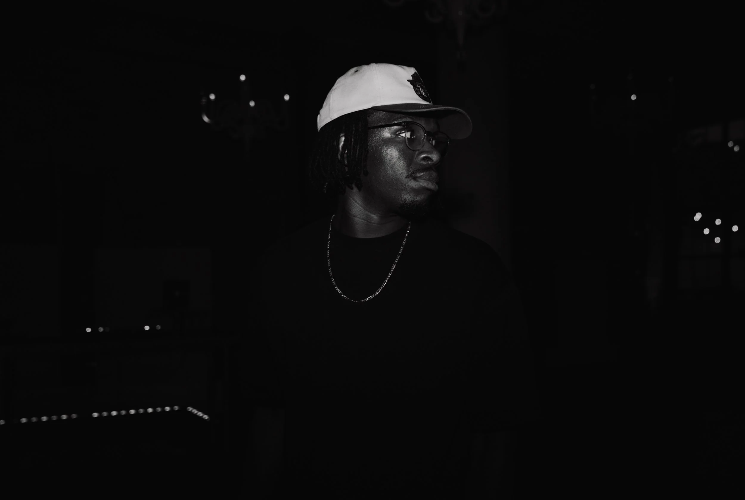 A person wearing glasses, a white cap, and a chain necklace, in a dark setting with some illuminated background lights.