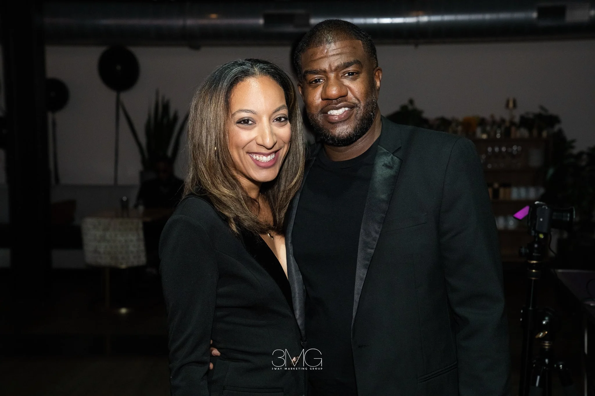 A woman and a man dressed in formal wear posing together at an indoor event. The woman has light brown hair and is wearing a black dress. The man has short hair, a beard, and is wearing a black tuxedo. Both are smiling and looking at the camera.