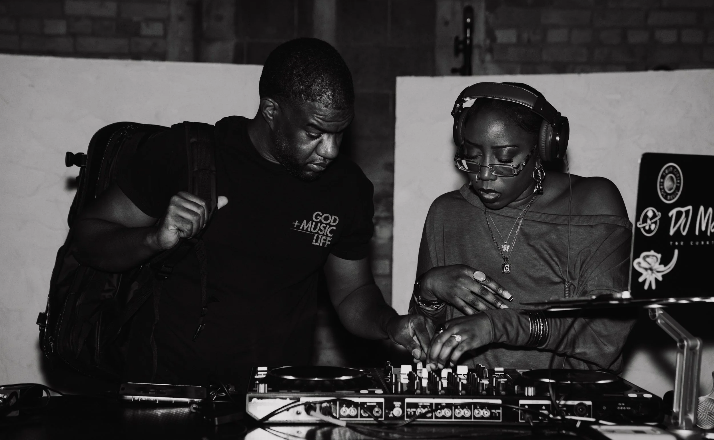 Two people working together at a DJ setup, one man and one woman, both focused on the equipment, with the woman wearing headphones and glasses, the man carrying a backpack, in a black and white photo.