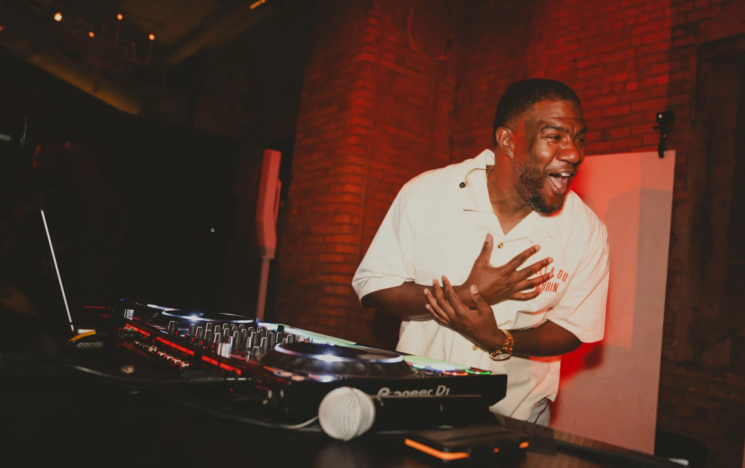A man djing at a venue with a red brick wall background, wearing a white shirt, and appearing animated and engaged with the music.