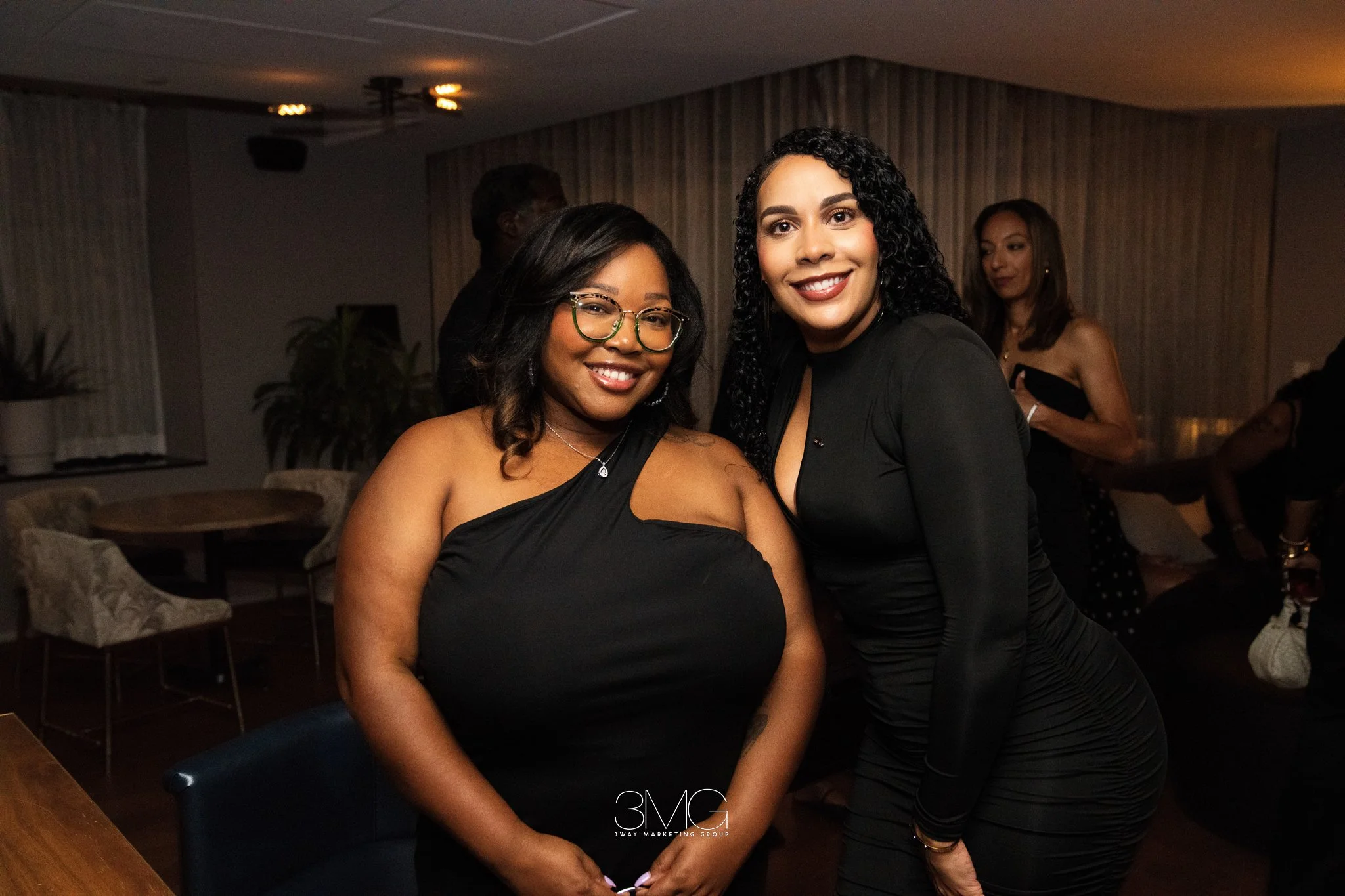 Two women smiling at a social gathering, one with glasses and the other with curly hair, wearing black dresses, inside a dimly lit room with wood-paneled walls and chairs.