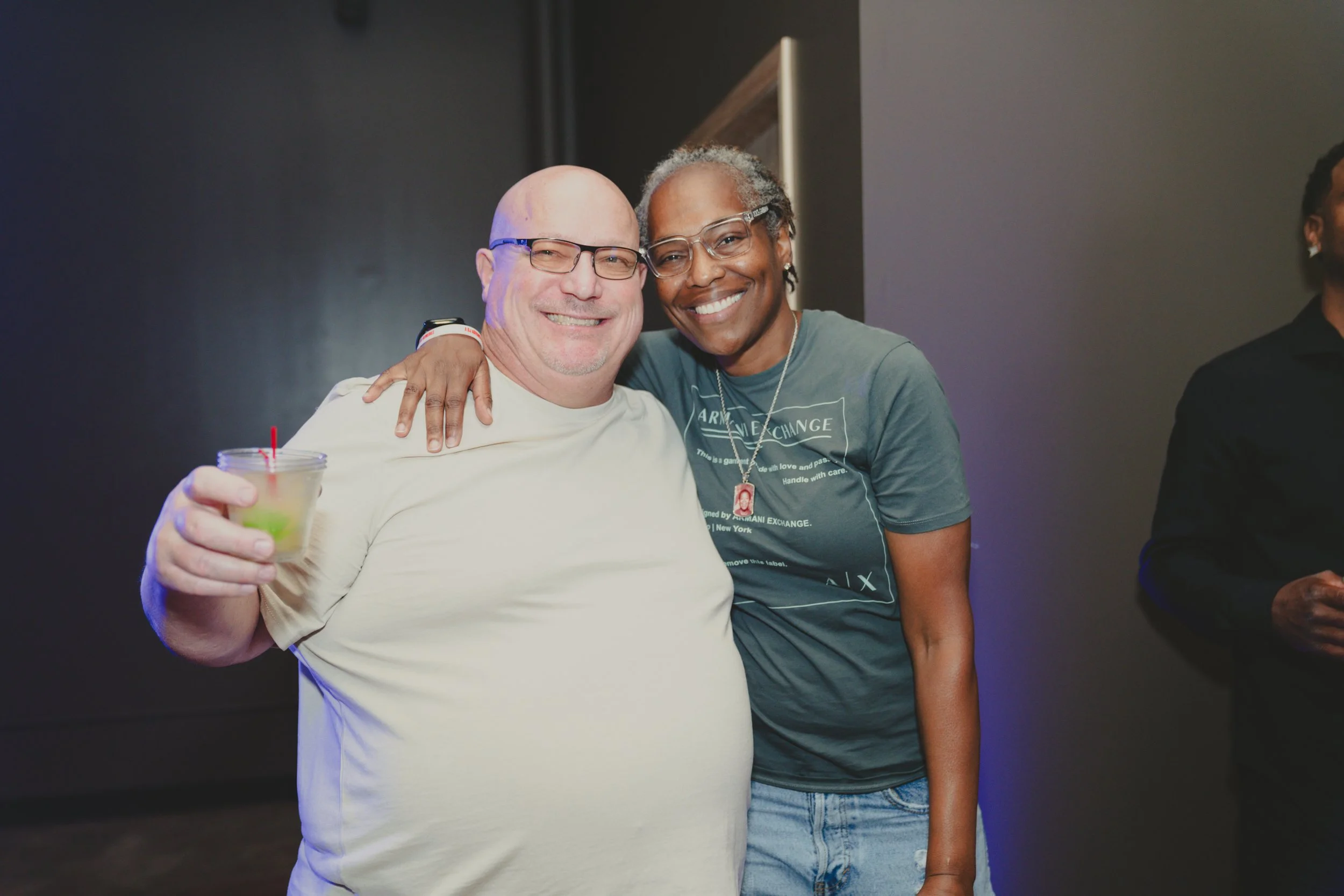 Two smiling people, a man with glasses holding a drink and a woman wearing glasses, hugging each other at a social event.