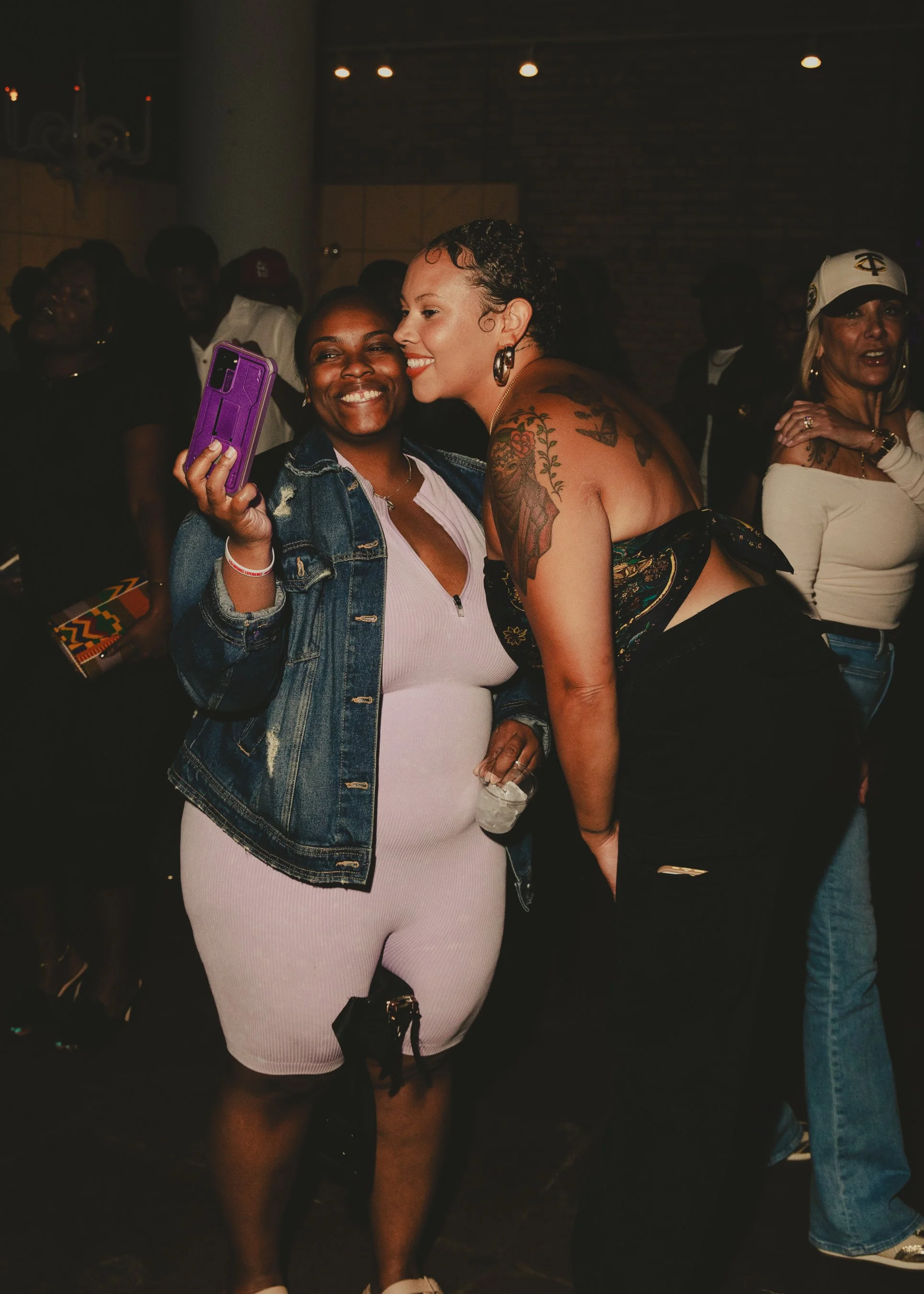 Two women smiling and taking a selfie in a dimly lit social setting with other people in the background.