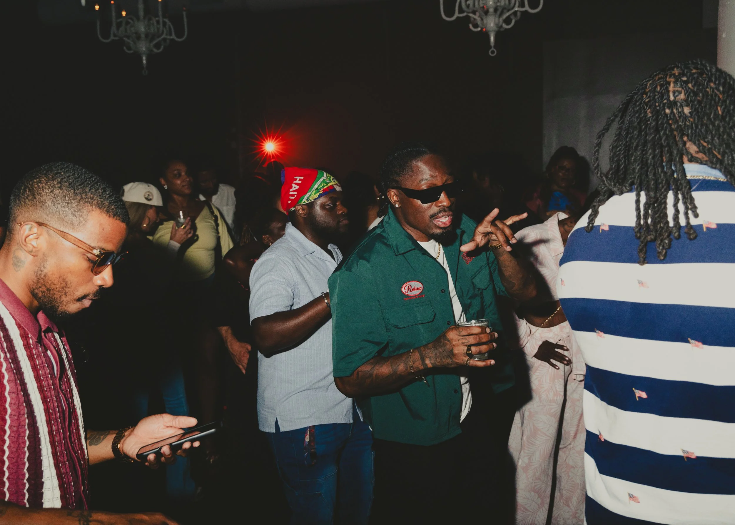 A group of people at a social gathering, chatting and using their phones, with one person in the center wearing sunglasses and a green shirt, and others in casual attire.