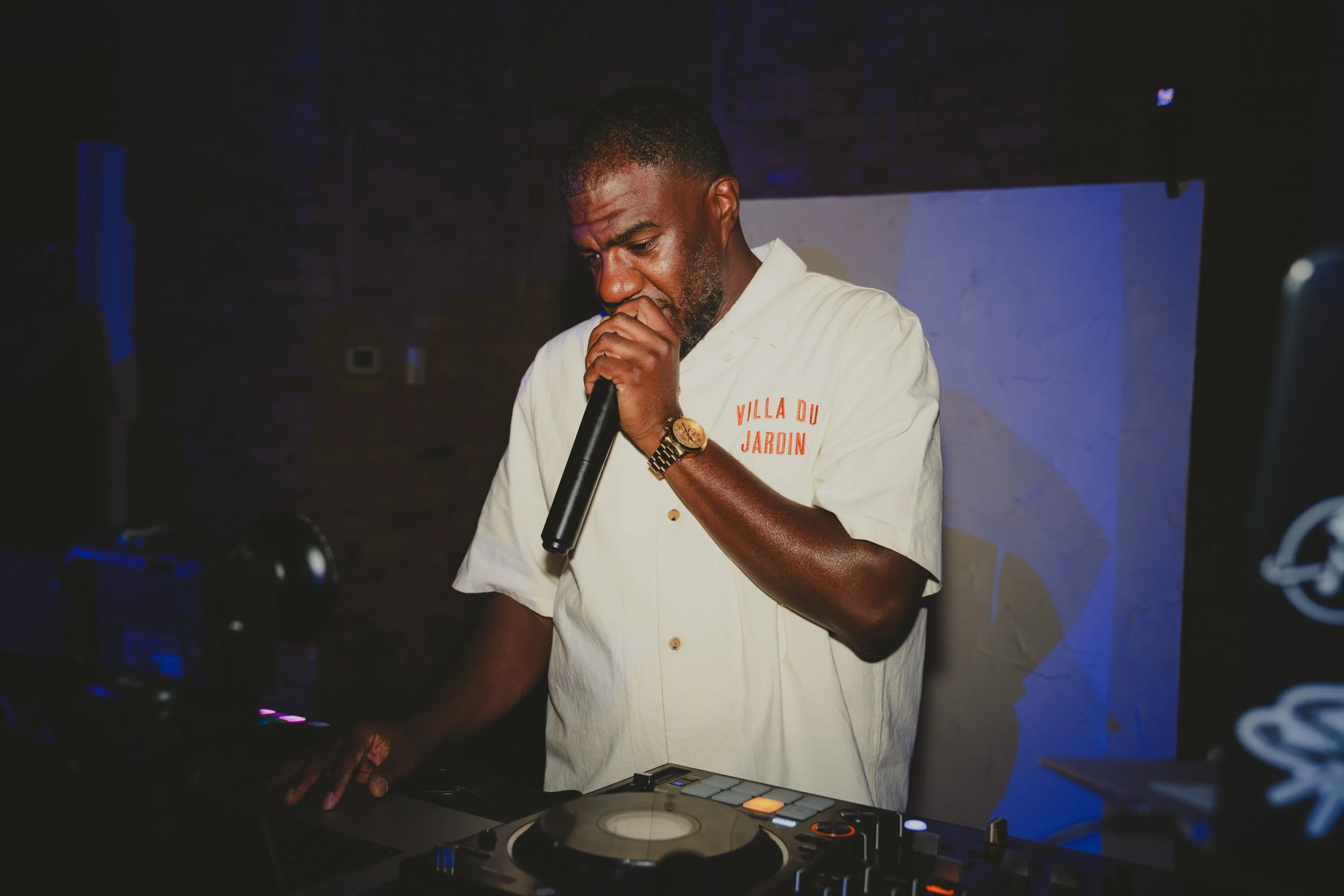A man in a white shirt with orange text 'Villa du Jardin' holding a microphone and DJ equipment, performing in a dark, possibly club or event setting.