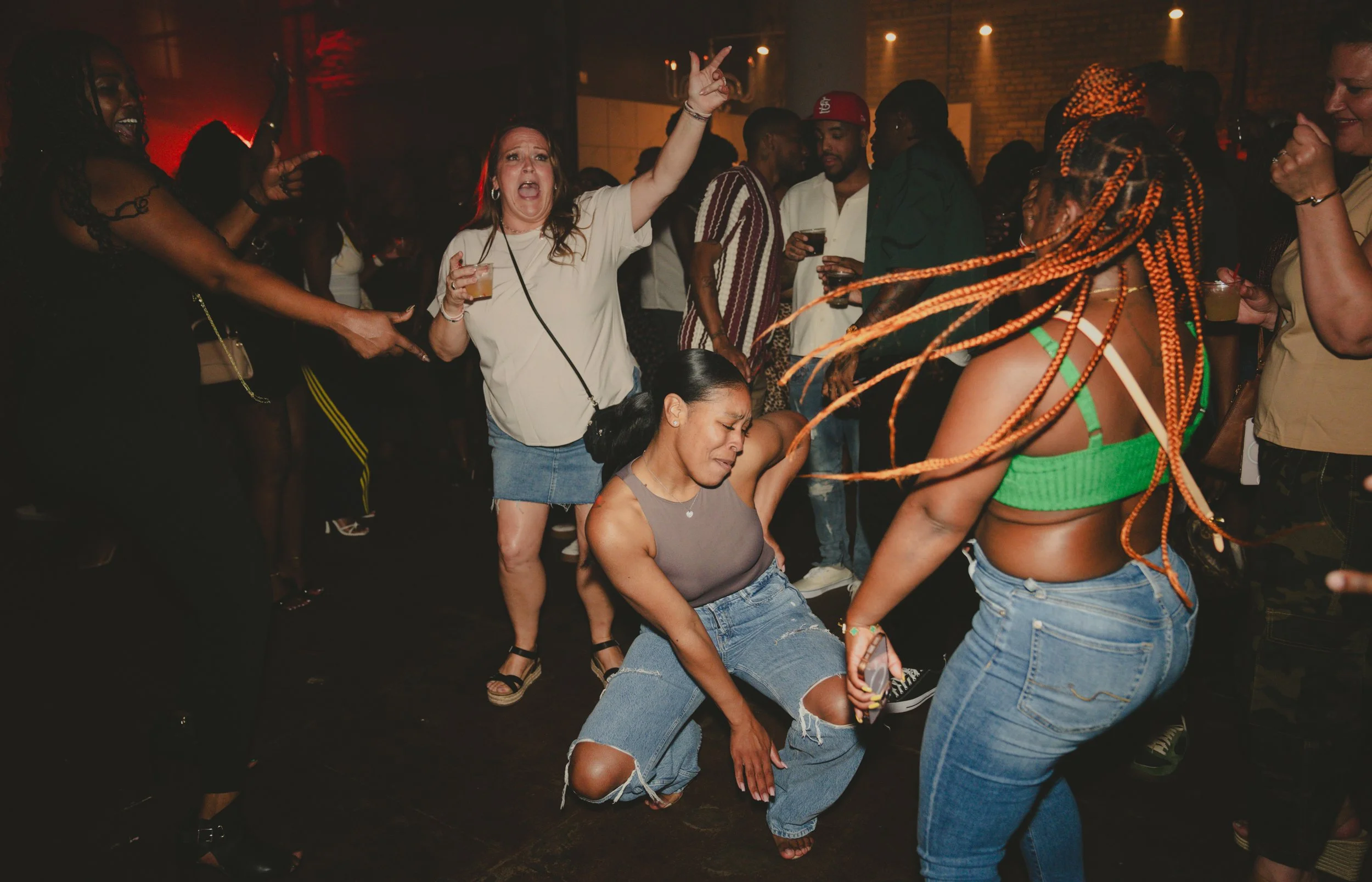 A group of people dancing and enjoying themselves at a lively indoor party, with some holding drinks and others with their hands in the air, in a dimly lit setting with warm lighting and brick walls.