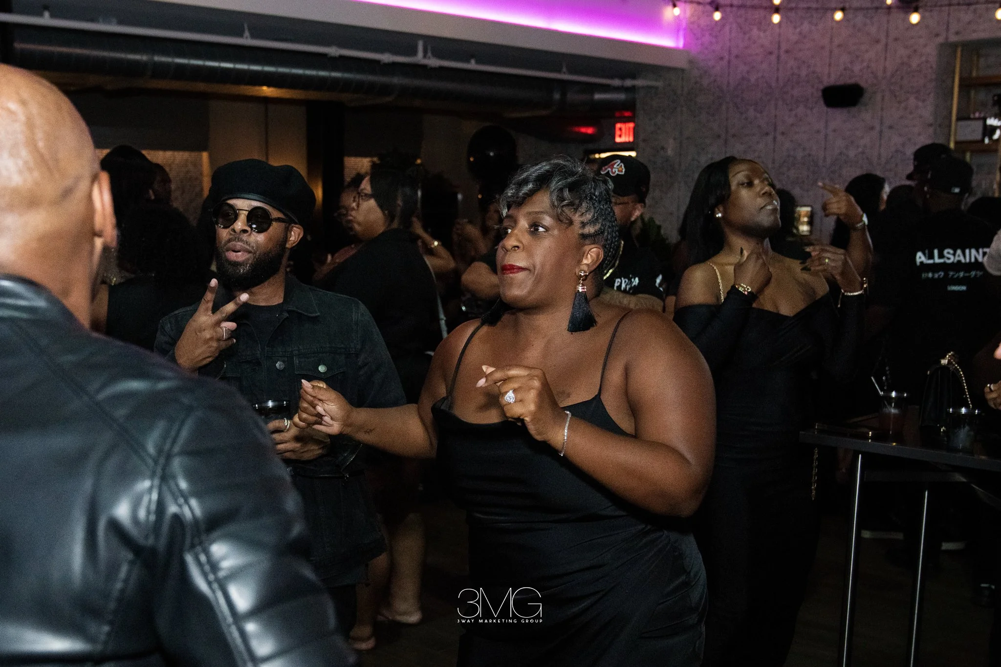 People socializing at a dimly lit indoor venue, including a man with sunglasses making a peace sign and a woman in a black dress with jewelry, surrounded by others engaged in conversation.