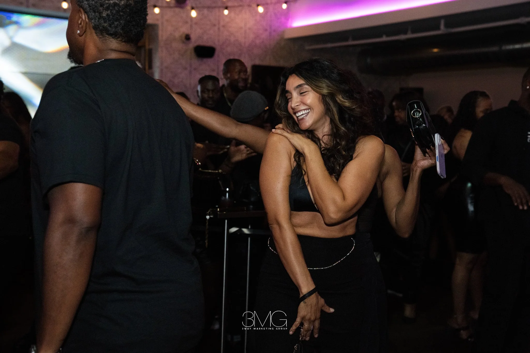 A woman with wavy hair and a black crop top smiling and dancing at a crowded party or club, with people in the background and purple and pink lighting.