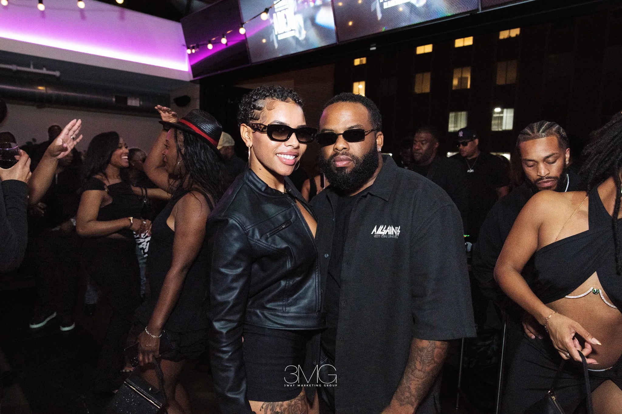 A group of people at a nightclub or party, with two people in the center wearing sunglasses, smiling and posing for the photo. The background shows other party attendees and colorful lighting.