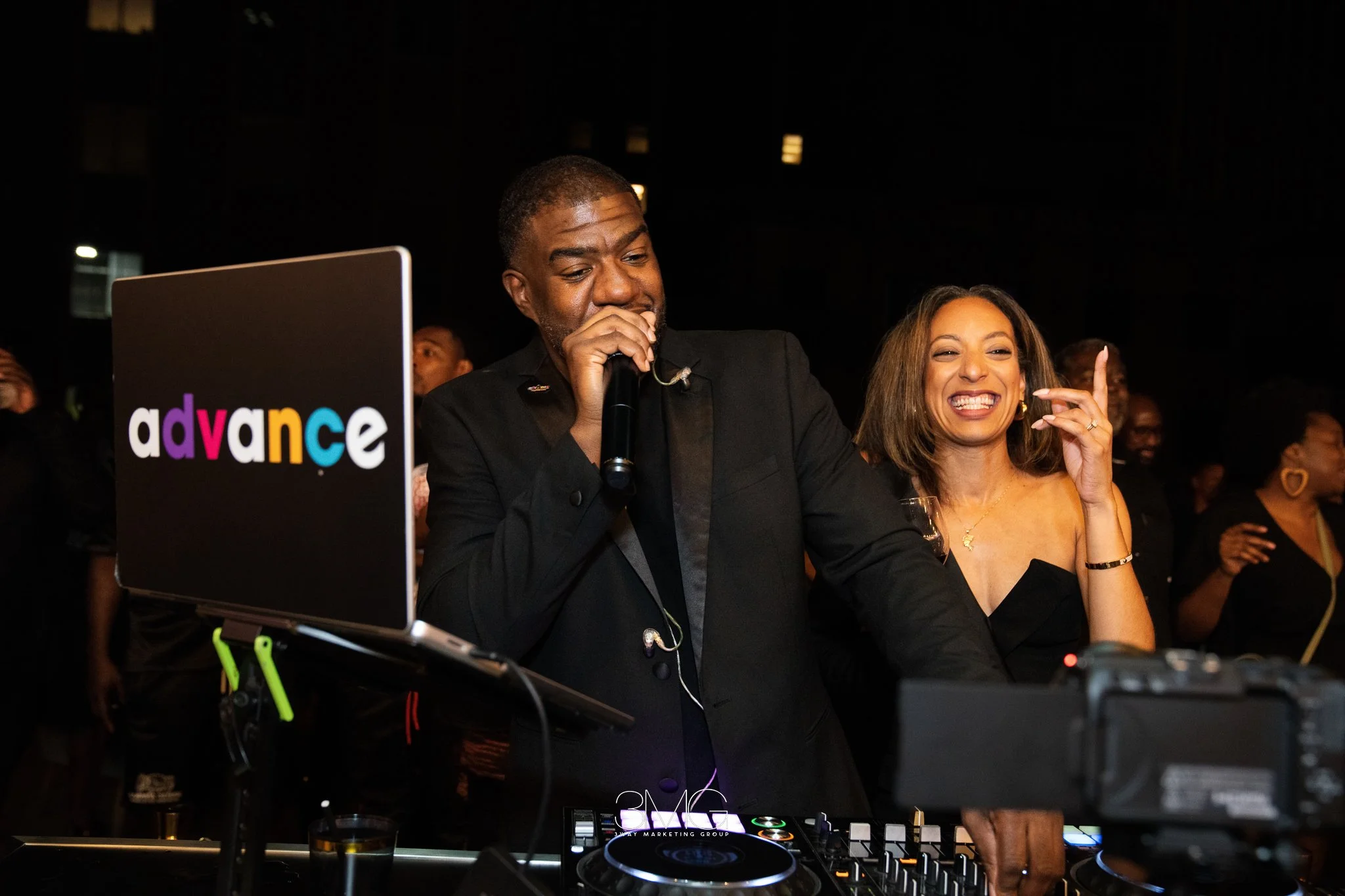 A man and woman dancing and smiling at a party, with DJ equipment in front of them. The woman is wearing a black dress and earrings, and the man is dressed in a black suit with a black shirt. A laptop with the word 'advance' is visible nearby, and th