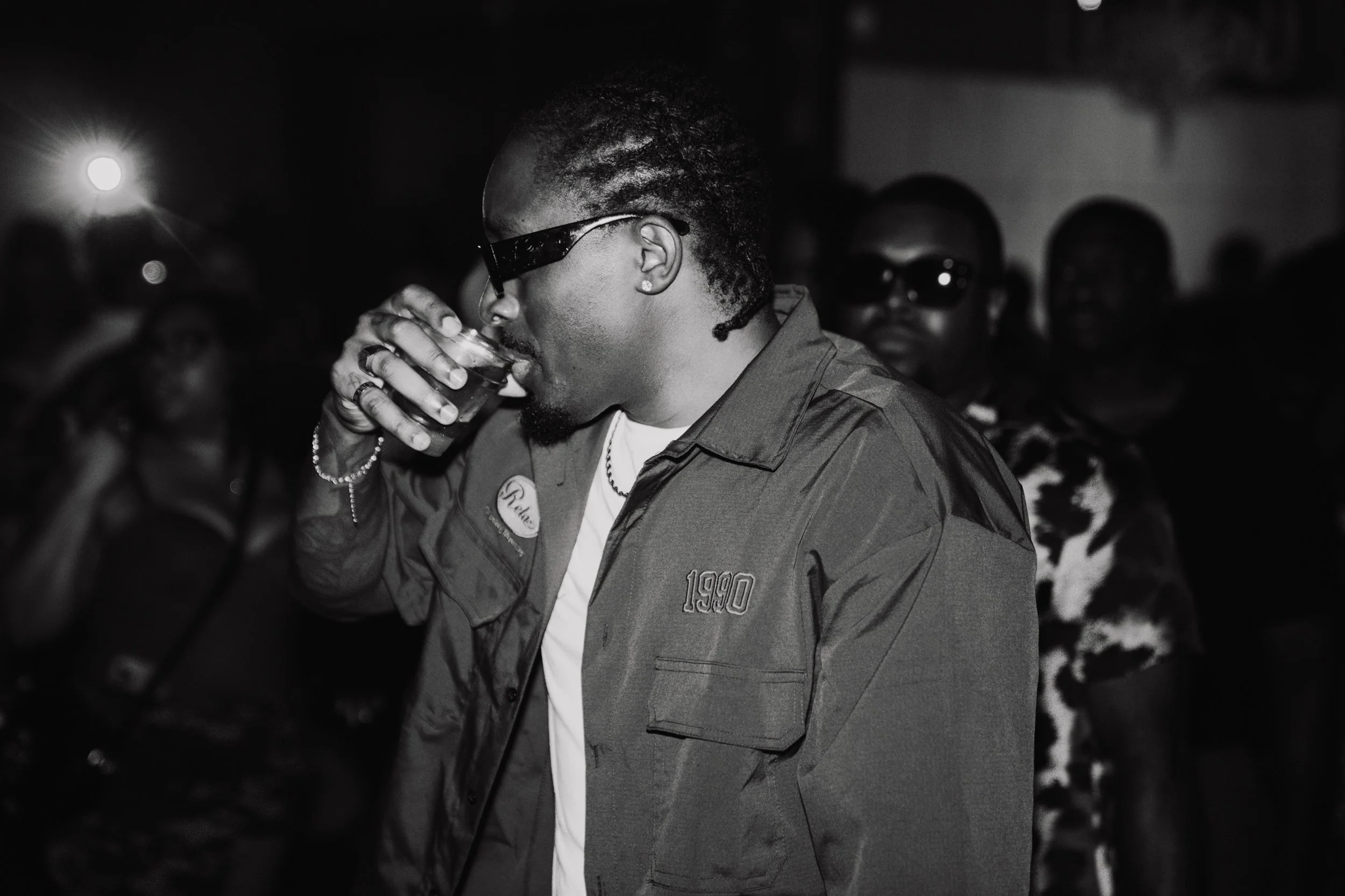 A man with glasses and a beard drinking from a glass at a social gathering at night, with other people in the background.