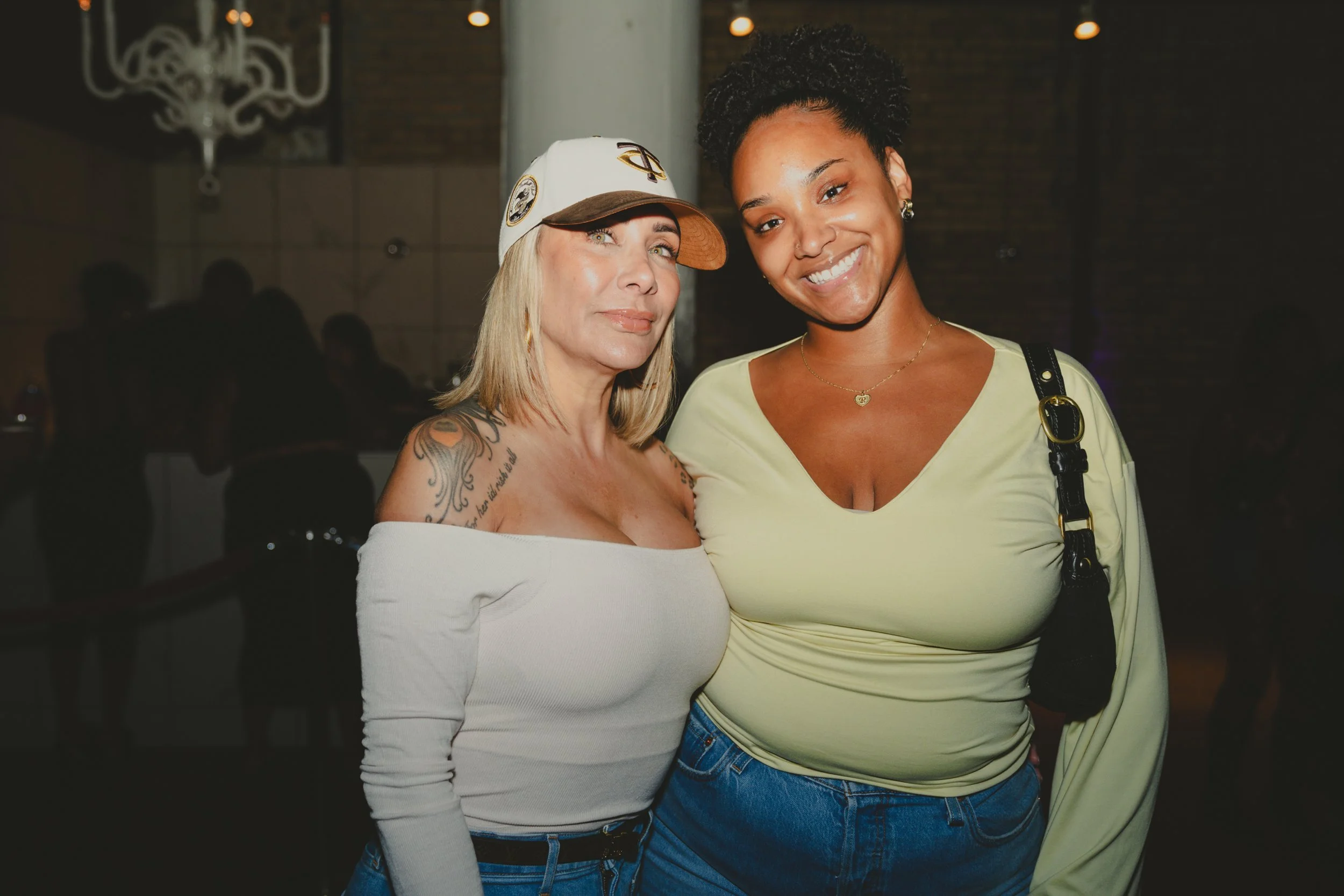 Two women smiling and posing together indoors, one with blonde hair and a white cap, the other with dark curly hair and a yellow shirt.