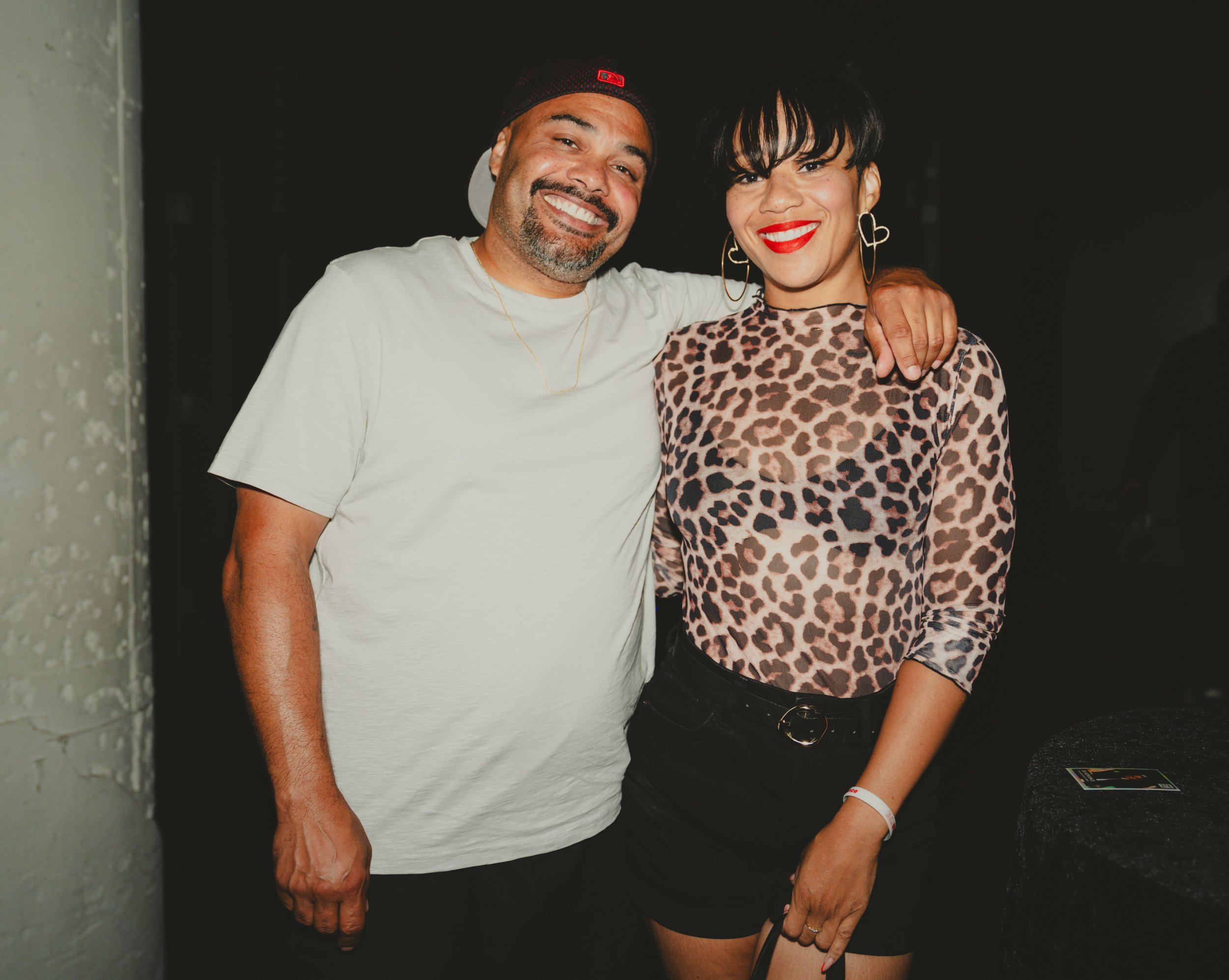 A man and woman smiling pose together at a social event, with the man's arm around the woman's shoulder. The man wears a black cap, white T-shirt, and has a beard, while the woman wears a leopard print top and red lipstick, with large hoop earrings a
