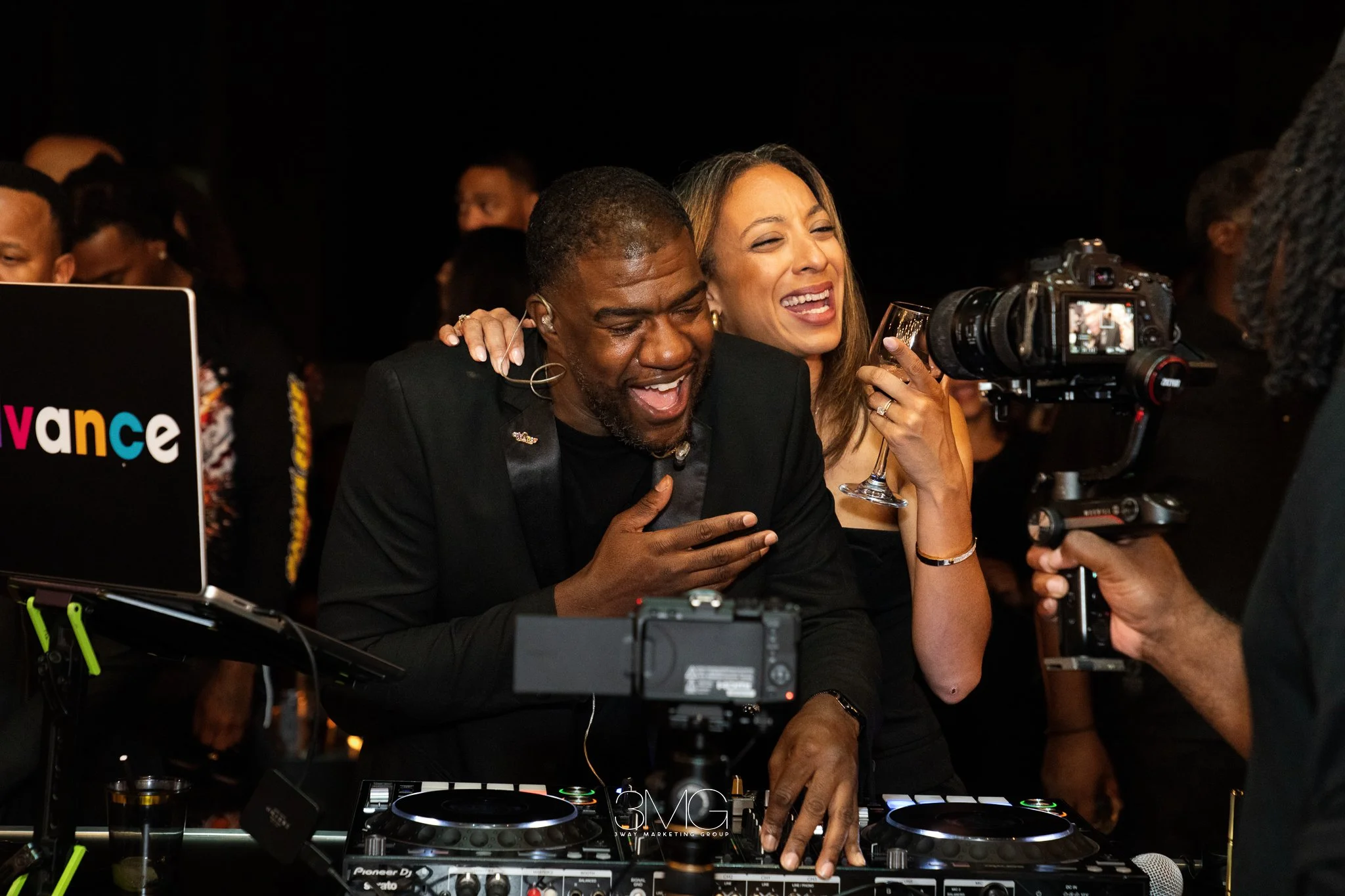 A man and woman laughing and enjoying themselves at a DJ event, with DJ equipment and a camera in the foreground.