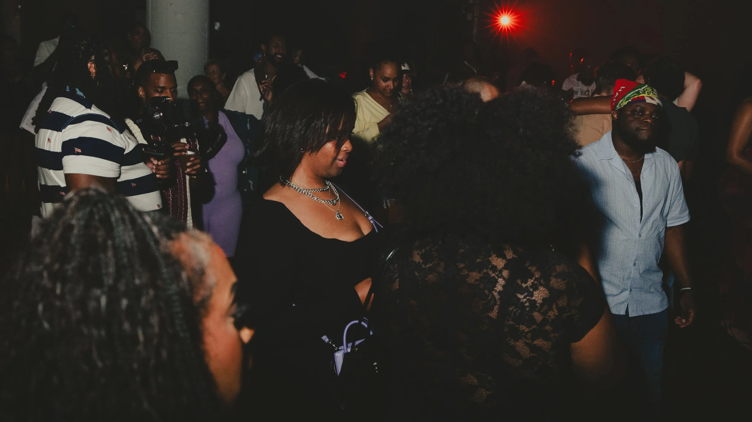Group of people at a party or concert in a dark indoor setting, some dancing and others watching, with a red lights flash in the background.