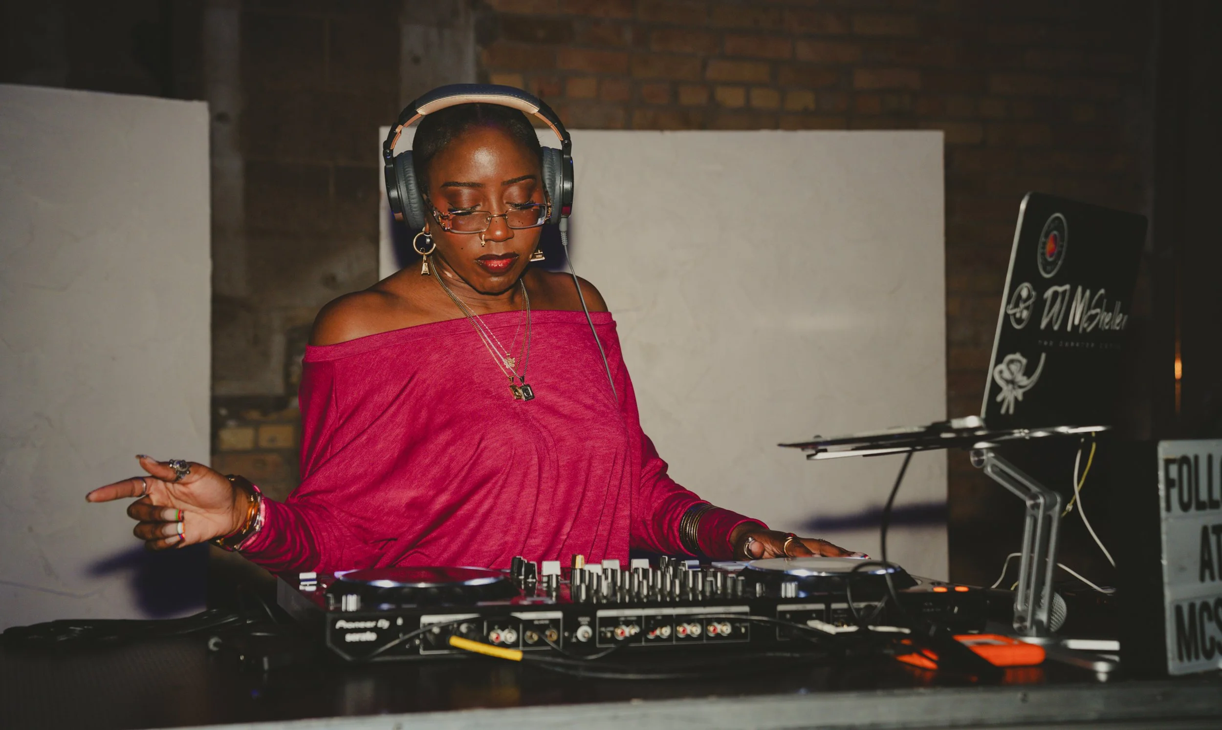 A woman with dark skin wearing a bright pink off-shoulder top, glasses, and headphones, is DJing behind music equipment in a dimly lit room with brick and concrete walls.