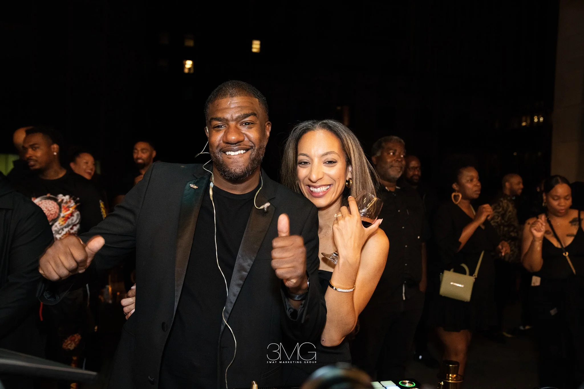 A man and woman smiling at a party, with the man giving a thumbs up. The man is dressed in a black suit, and the woman is holding a glass of wine. Other people are visible in the background enjoying the event.