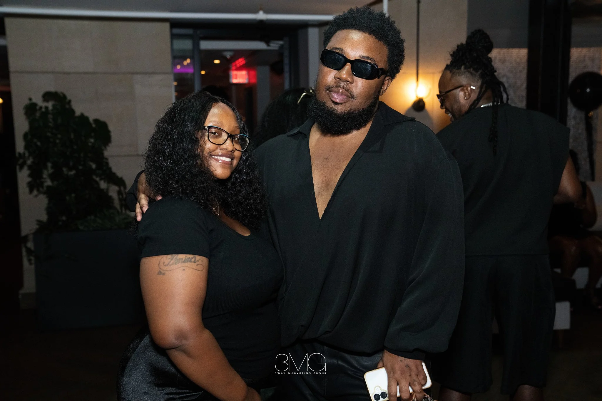 A man wearing black sunglasses and a black shirt, standing next to a woman with glasses, curly hair, and a tattoo on her arm. They are at an indoor event with dim lighting, smiling and posing for the camera.