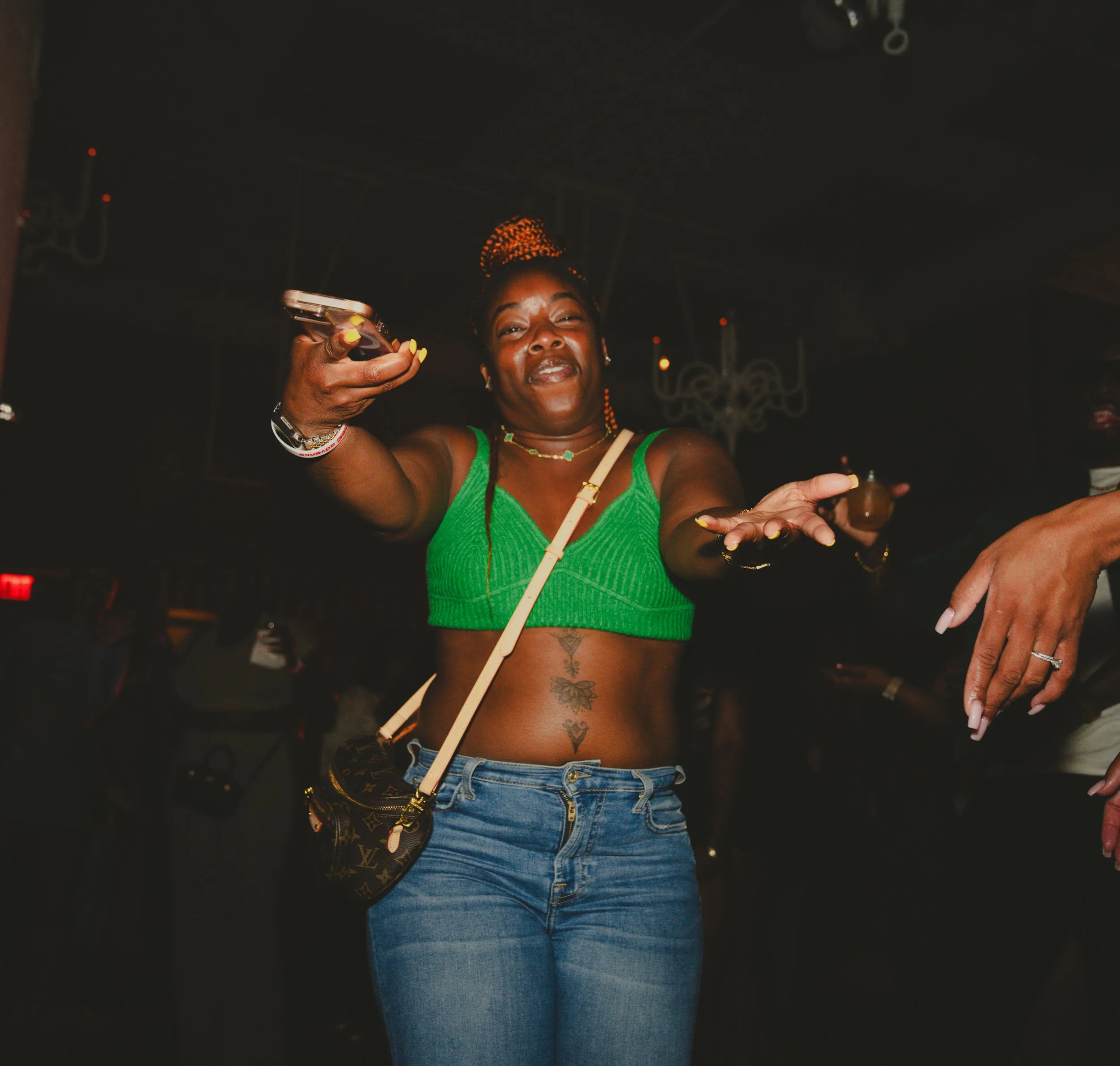 A woman with a plant-like hairdo, wearing a green sleeveless top and blue jeans, dancing at a party or nightclub with a dark background.