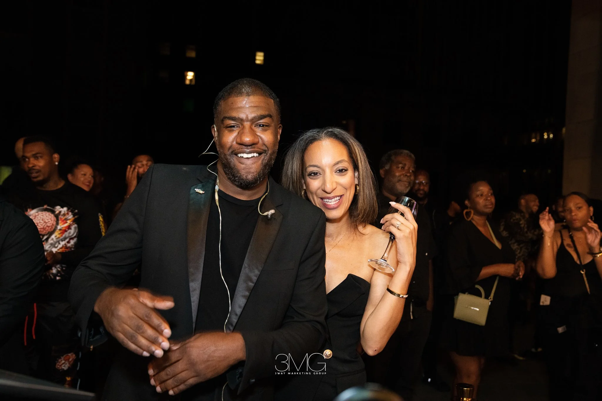 A group of people at a social event, with a man and woman in the foreground smiling and holding drinks, others in the background socializing at night.