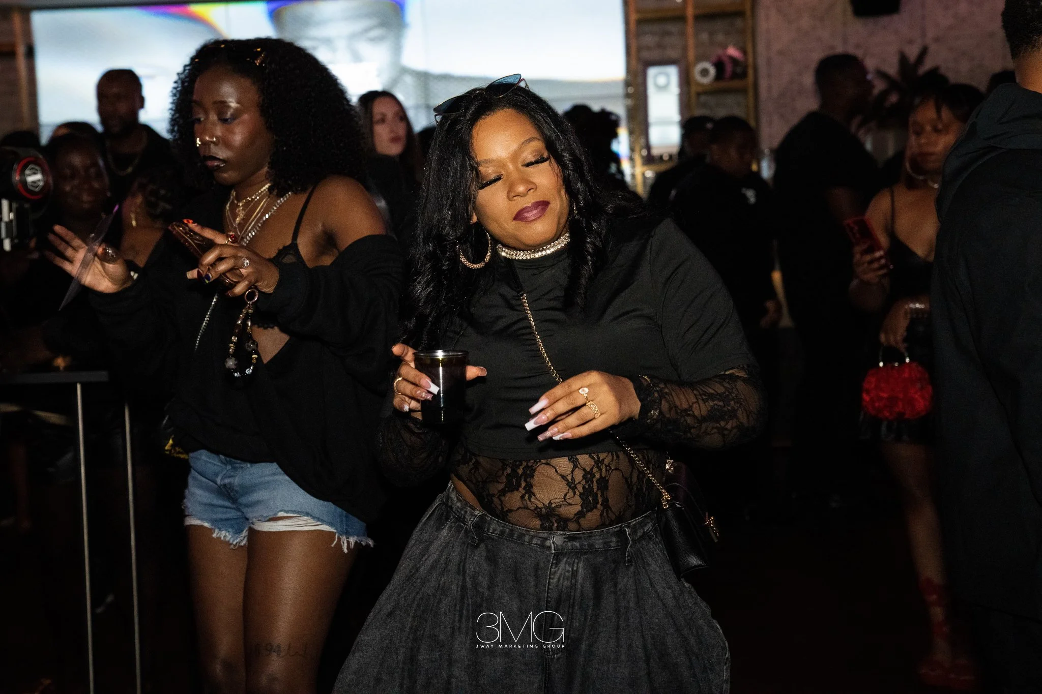 A woman with long black hair wearing a black lace top, jewelry, and sunglasses on her head, is in a crowd at a dimly lit indoor event, holding a drink with other people surrounding her.