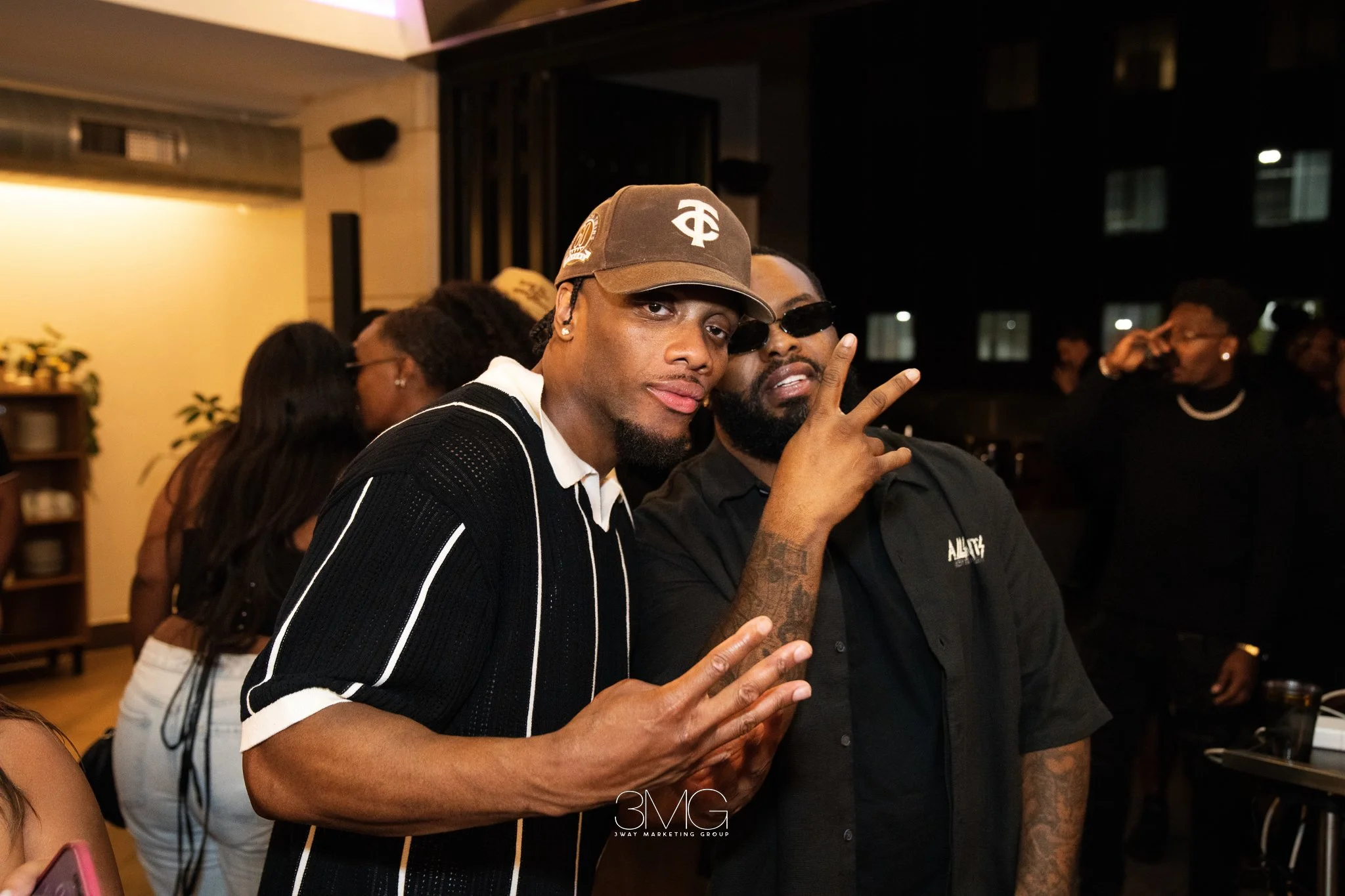 Two men posing at an indoor event, one making a peace sign, other with hand near face, other people in background, warm lighting.