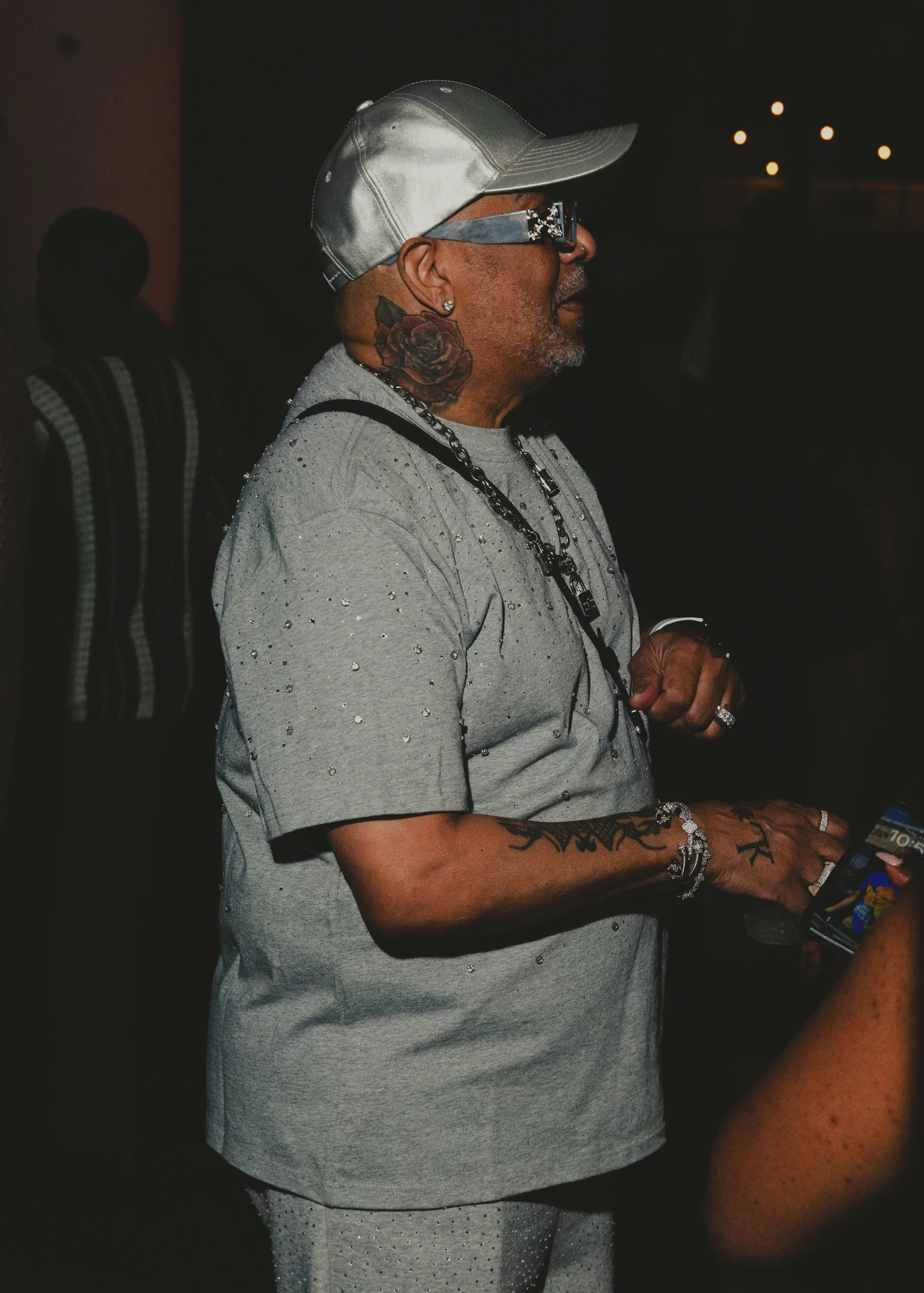 A man wearing a silver cap, large glasses, and a light grey shirt with rhinestones. He has tattoos on his neck and arm, and is holding a smartphone. The background is dark with a few blurred lights.