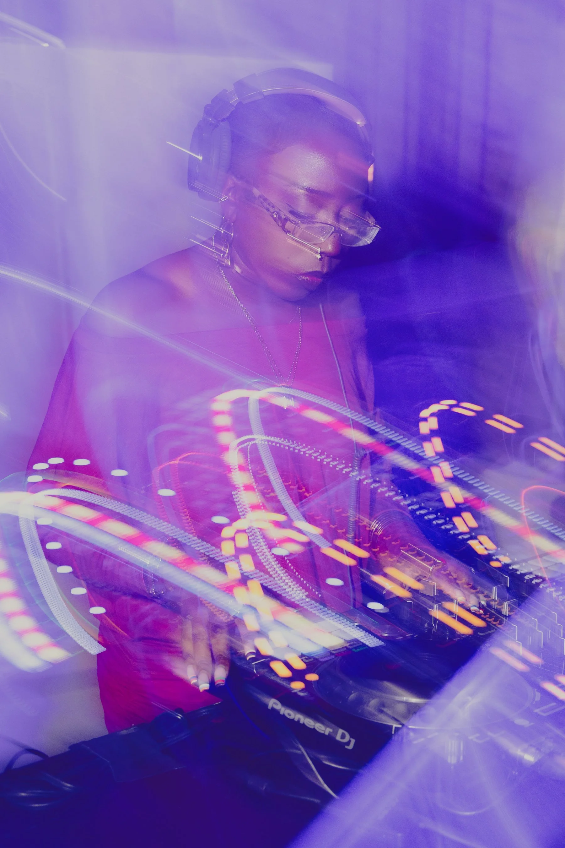 A woman with dark hair and glasses operating a DJ controller with colorful light effects and a purple haze.
