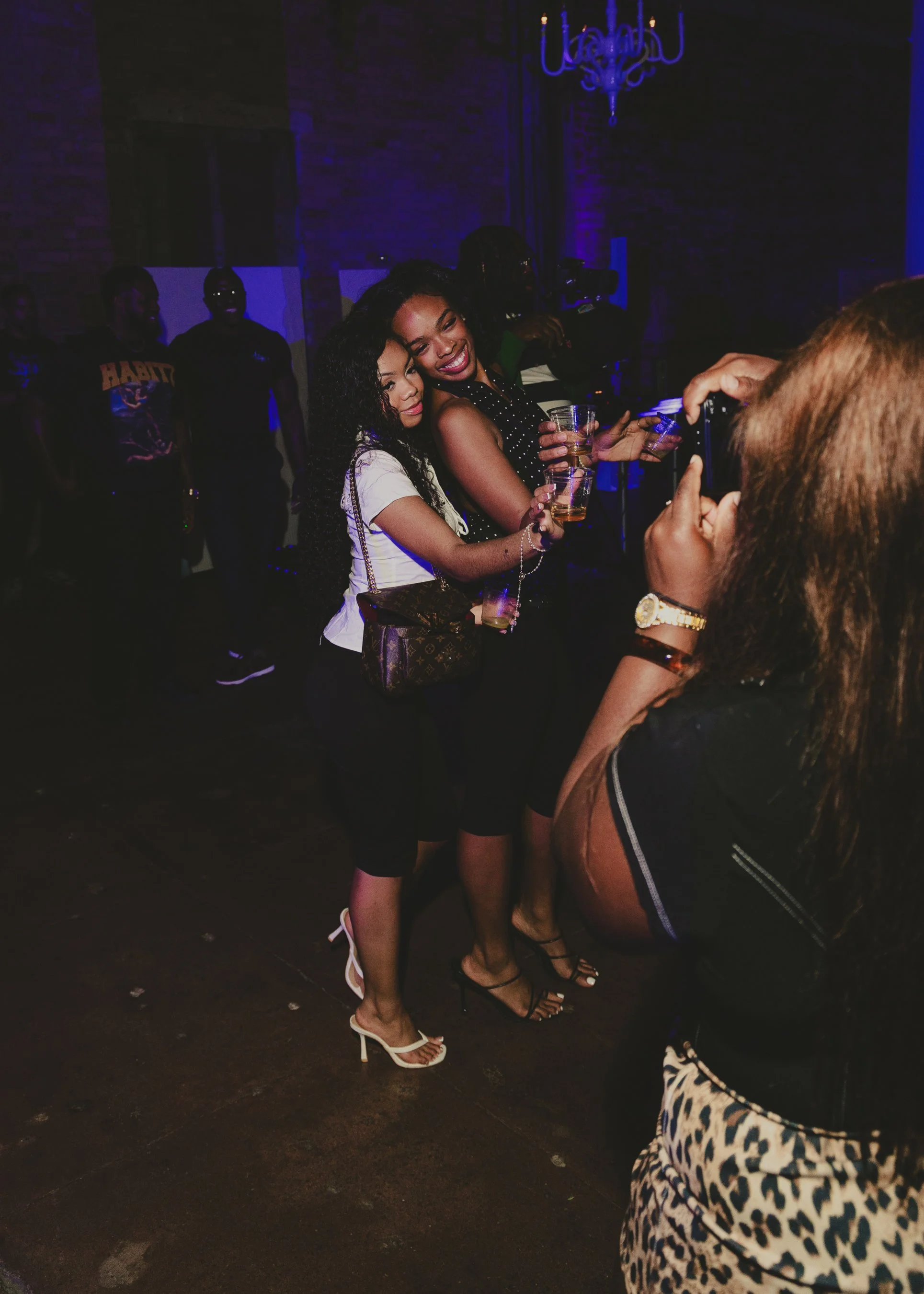 Three women smiling and posing for a photo at a party or nightclub, holding drinks. The background is dark with blue lighting, and other people are visible in the background.