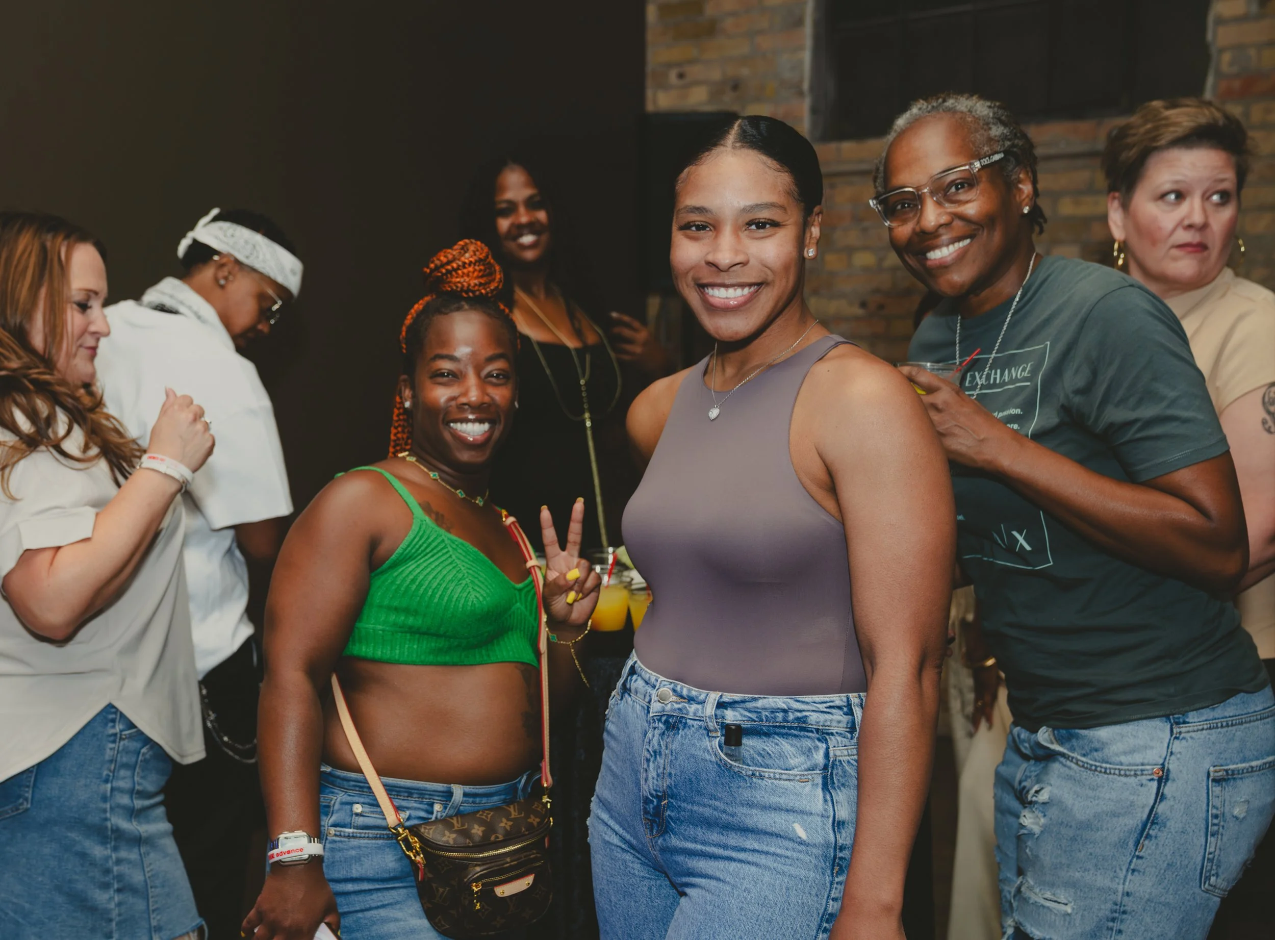 A group of smiling women at a social gathering, standing close together in front of a brick wall.