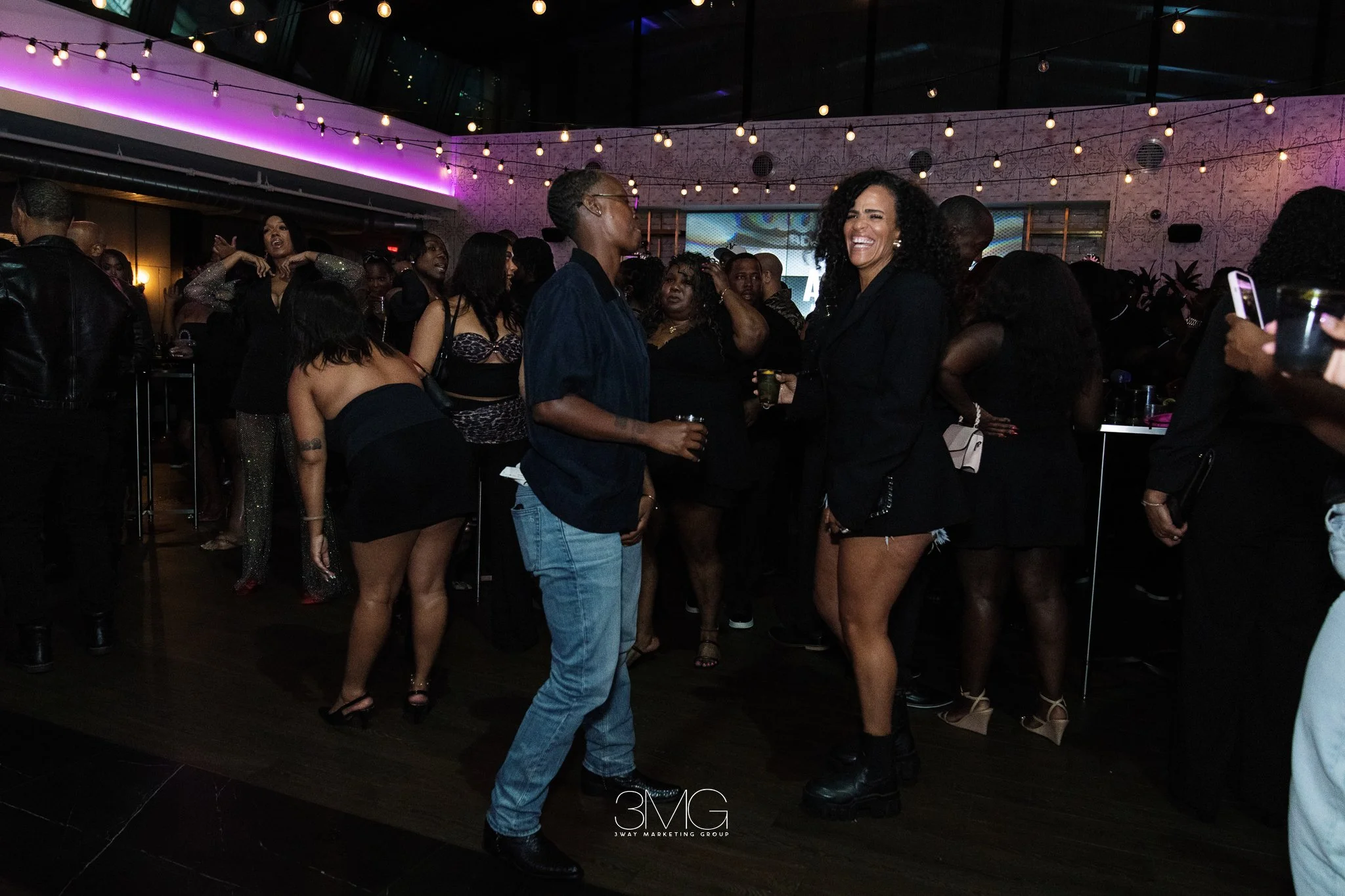 People dancing and socializing at a party with string lights and colorful lighting, including women in fashionable outfits and a woman with curly hair smiling.