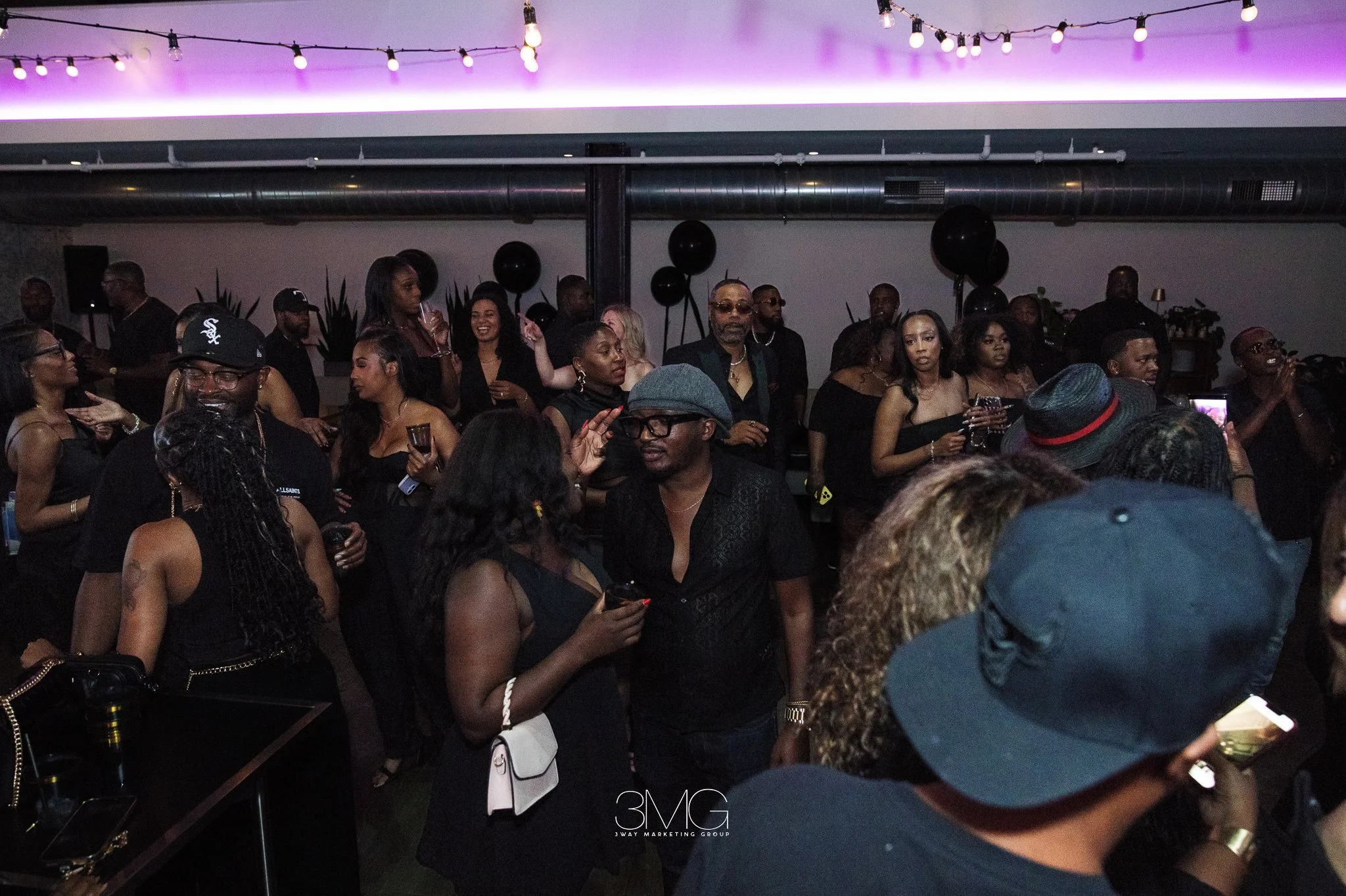 A crowded indoor party with people dancing and socializing, decorated with black balloons and string lights, with purple ambient lighting.