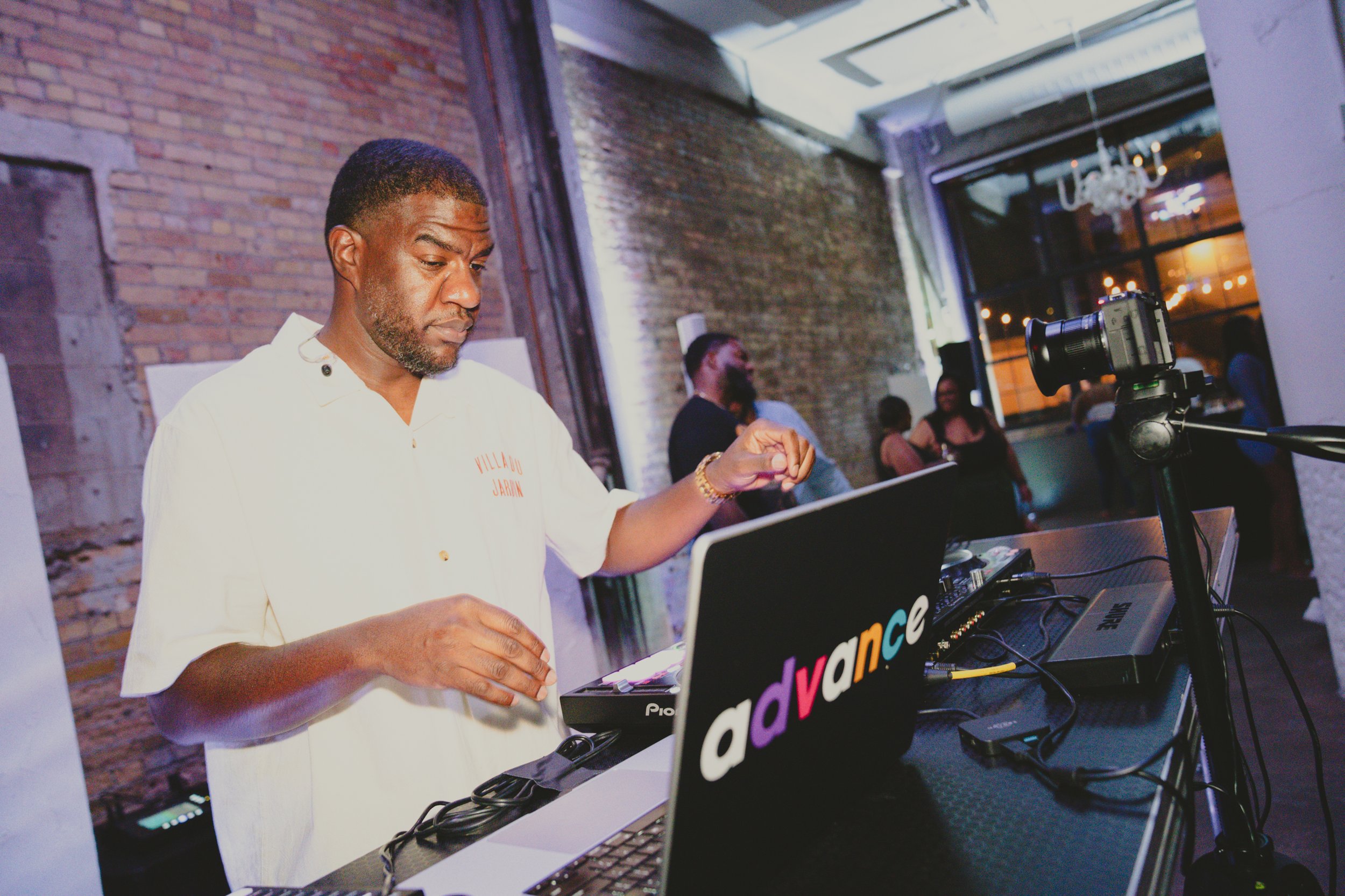 A DJ is performing at an indoor event with a brick wall and a large window in the background. The DJ is wearing a white shirt and is focused on his equipment, which includes a laptop and a DJ controller with colorful branding. A camera is set up on a