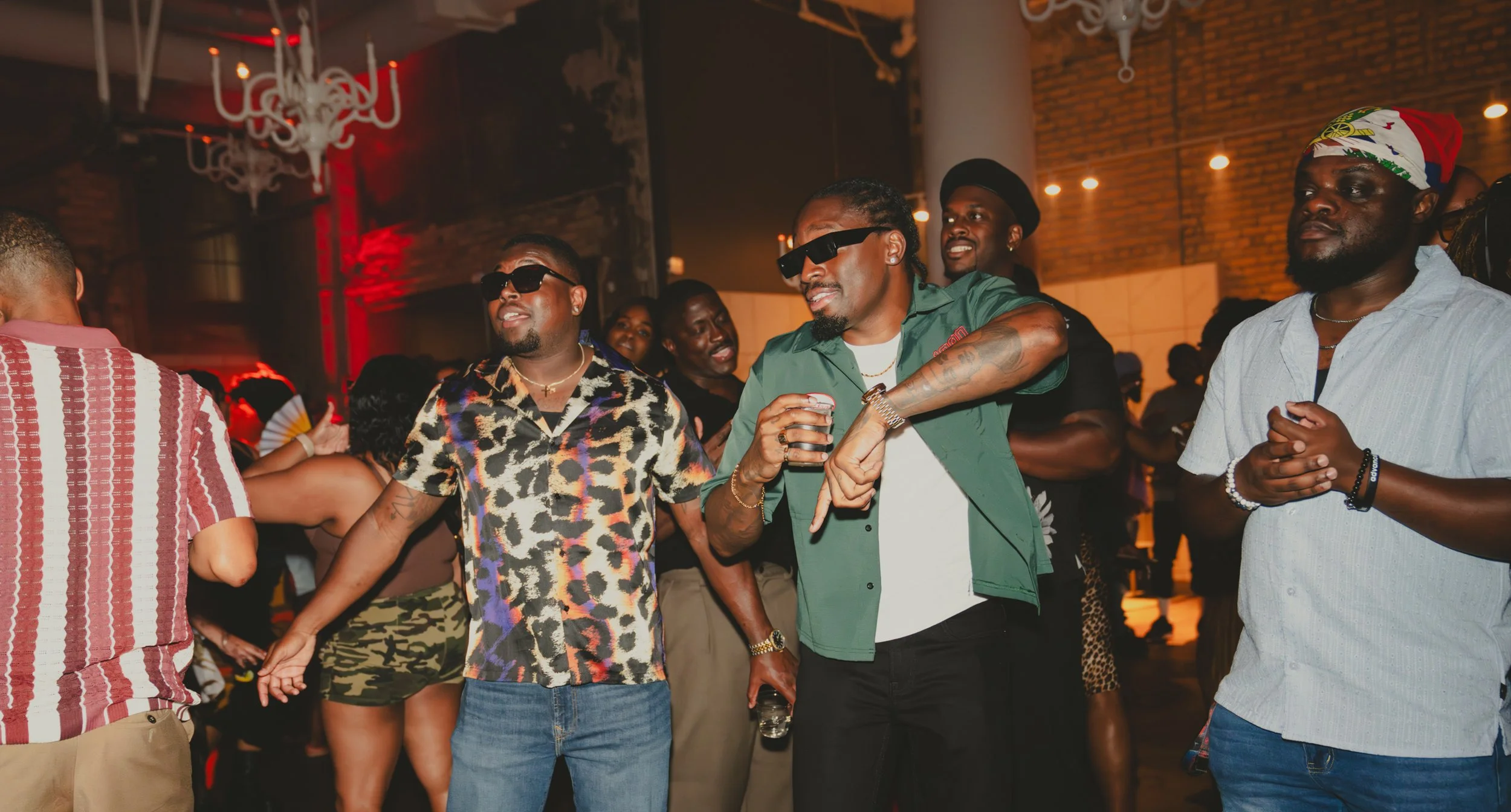 Group of people socializing at a party or club, some wearing sunglasses, colorful clothing, and accessories, with dim lighting and brick wall background.