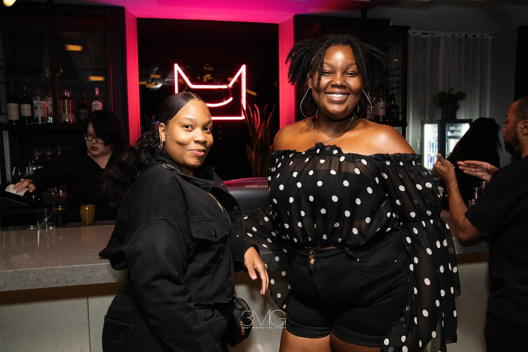 Two women smiling at a bar with a neon cat face sign in the background. The woman on the left has dark hair, is wearing a black denim jacket, and has her hand on her hip. The woman on the right has curly hair, is wearing a black off-the-shoulder polk