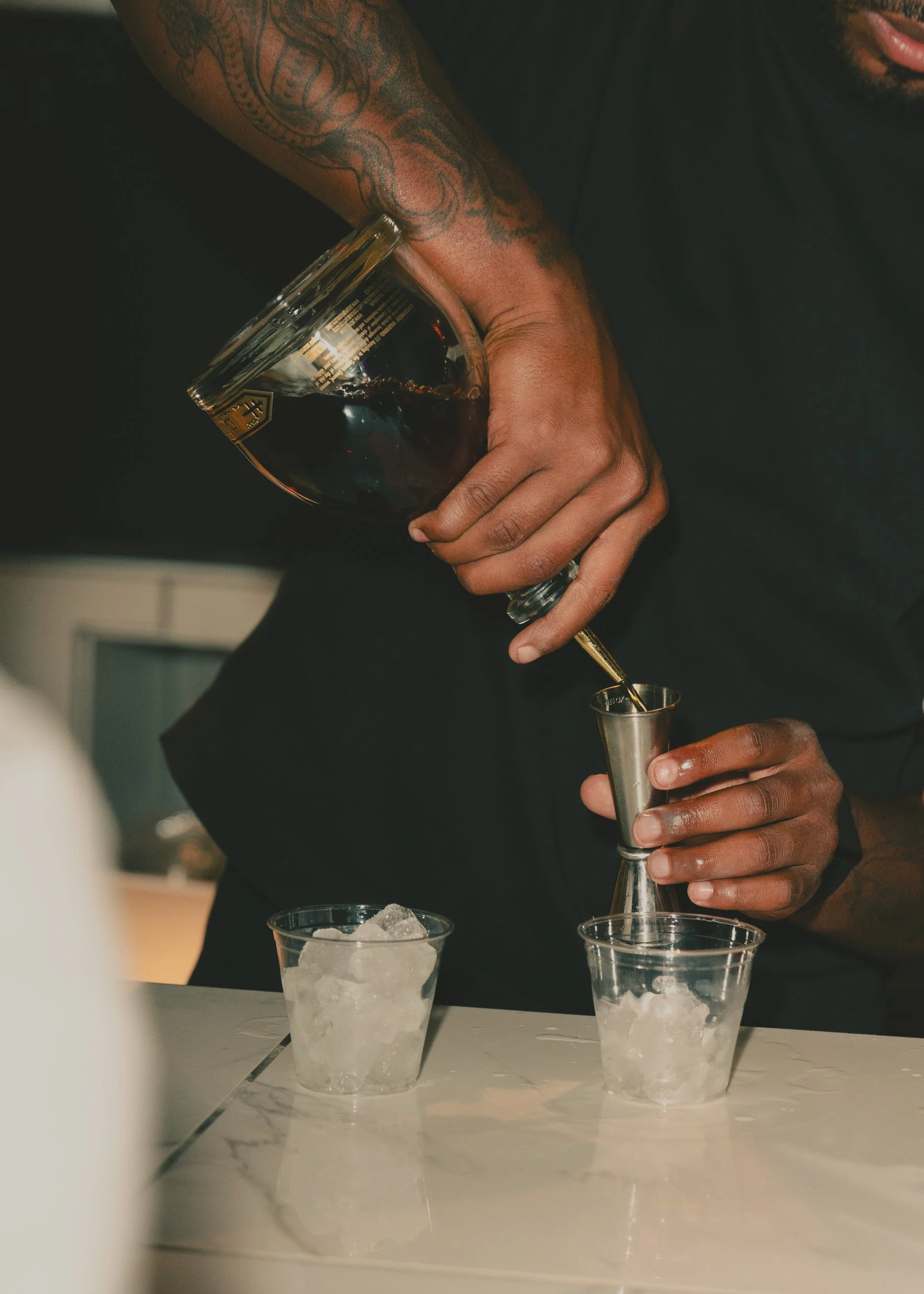 A person with tattoos on their arm pouring a dark liquid from a bottle into a jigger, with three glasses of ice and a glass container on a white marble countertop.