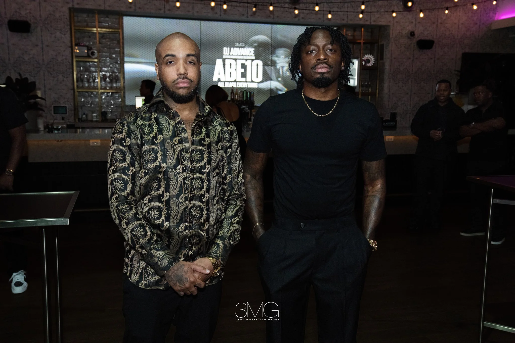 Two men standing side by side at an event in a bar or lounge area with a large screen behind them displaying the text "3MG ABEO ALL BLACK EVENTS". The man on the left has a bald head and beard, wearing a patterned shirt, with his hands clasped in fro