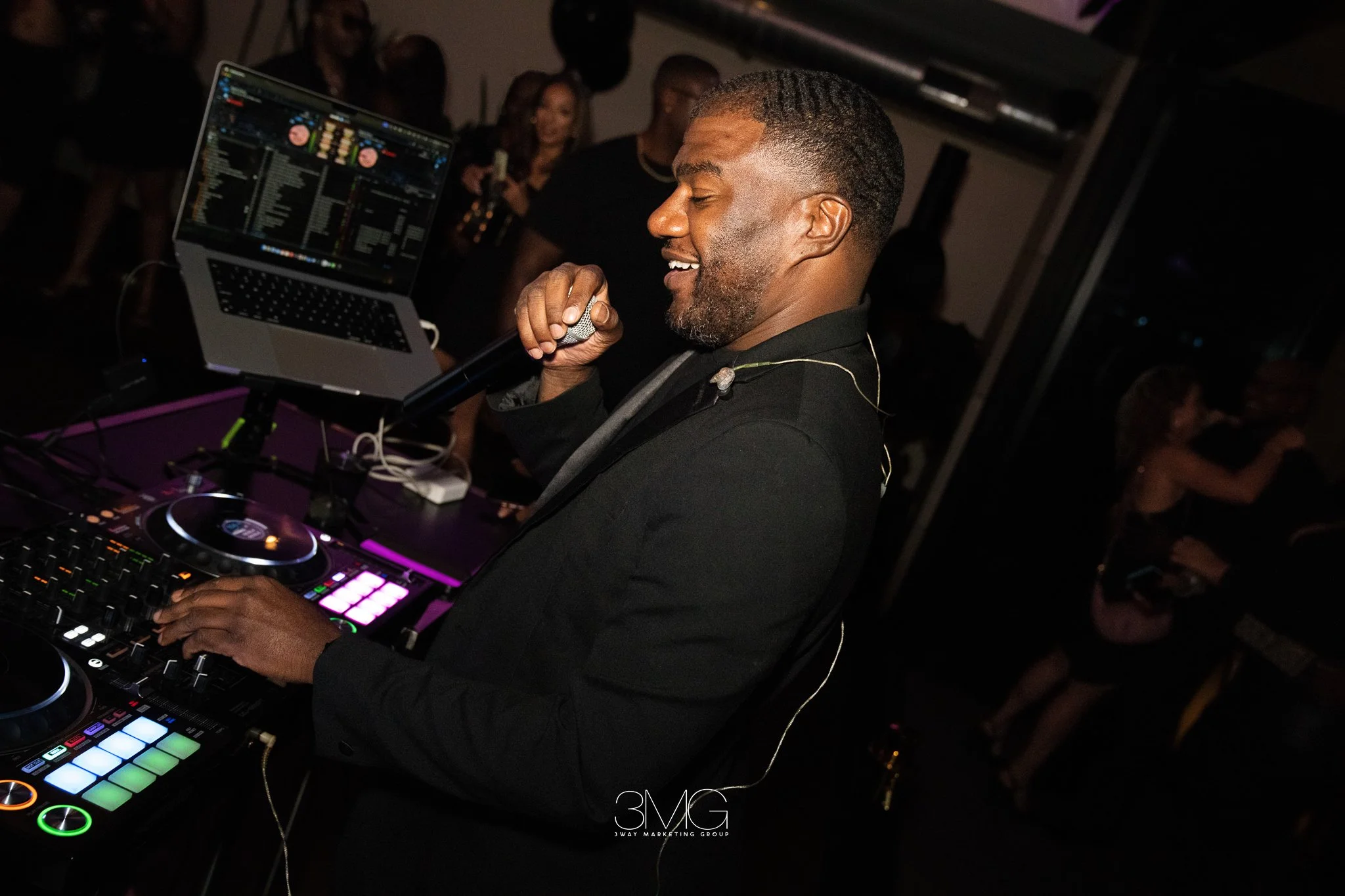 A man with short hair and a beard smiling while holding a microphone in front of DJ equipment in a dark club. There are people in the background, some holding drinks, with purple and black lighting.