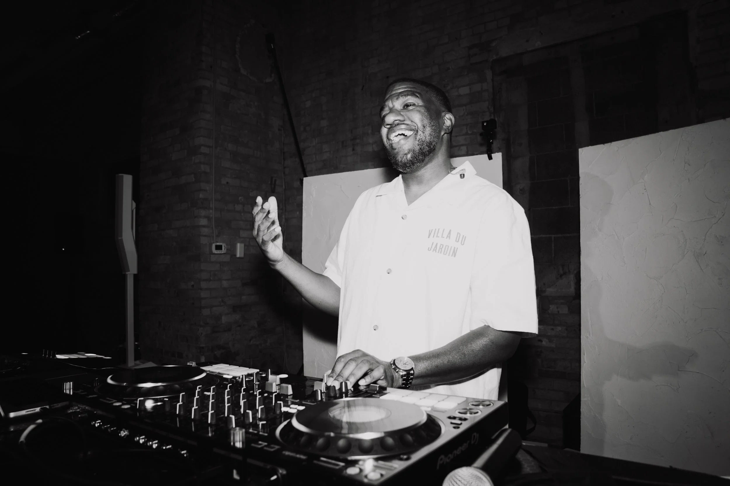 A man with a beard and short hair, wearing a white shirt with the text 'VILLA DU JARDIN,' is DJing with a DJ controller in front of him, smiling and dressed casually, in a dimly lit room with brick walls.