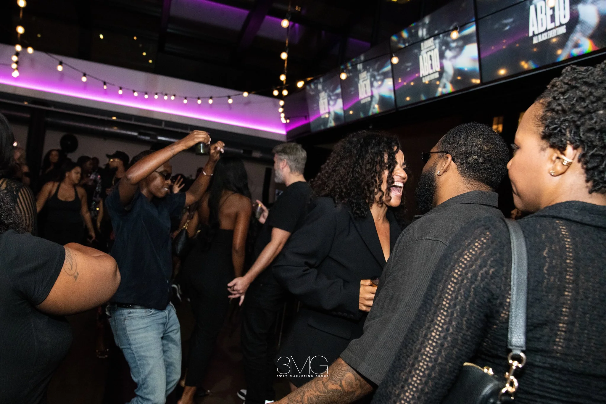 A diverse group of people dancing and socializing in a nightclub with dark lighting and colorful LED screens displaying promotional content for an event called 'ABELTO'. The atmosphere is lively and energetic.