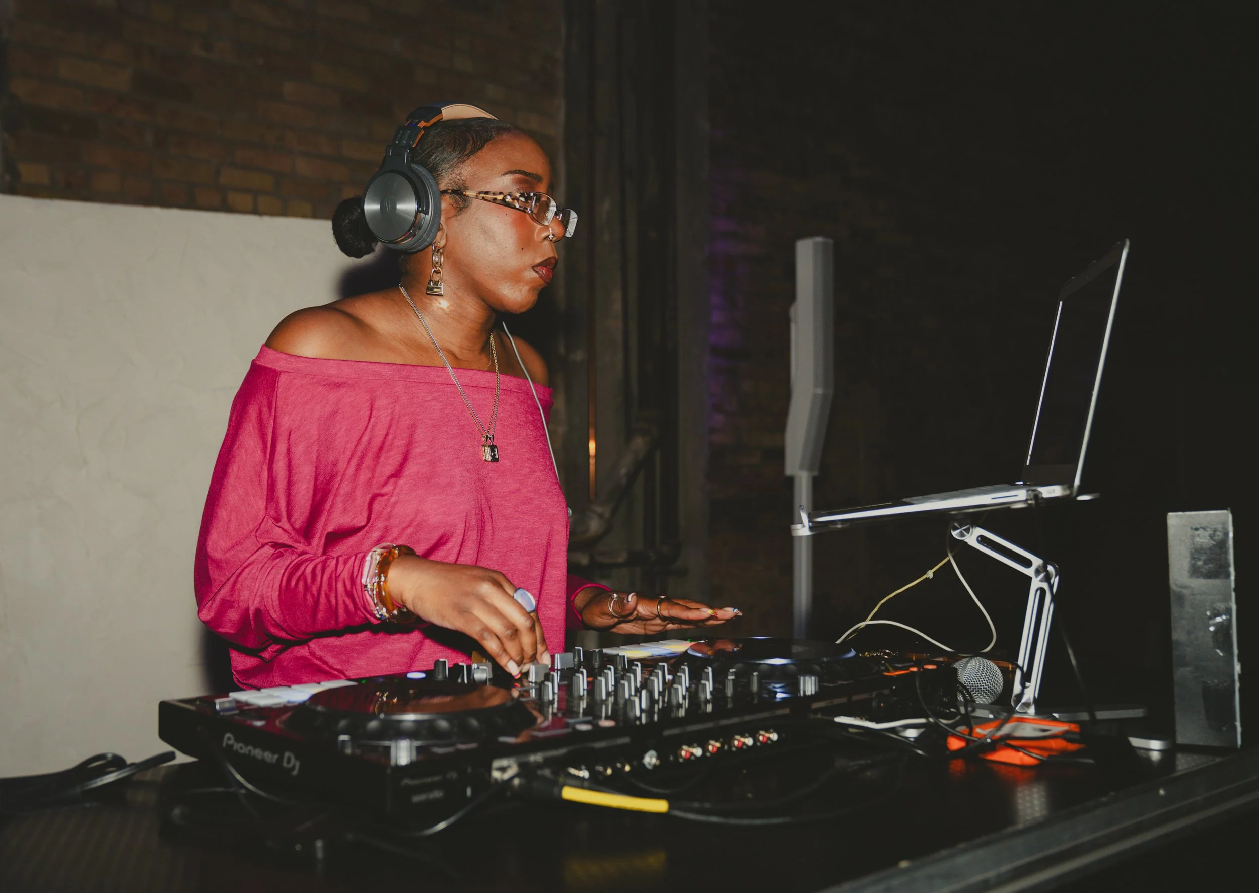 A woman DJ wearing a pink off-shoulder top, sunglasses, and large headphones, operating turntables and a laptop in a dimly lit indoor setting with exposed brick wall.