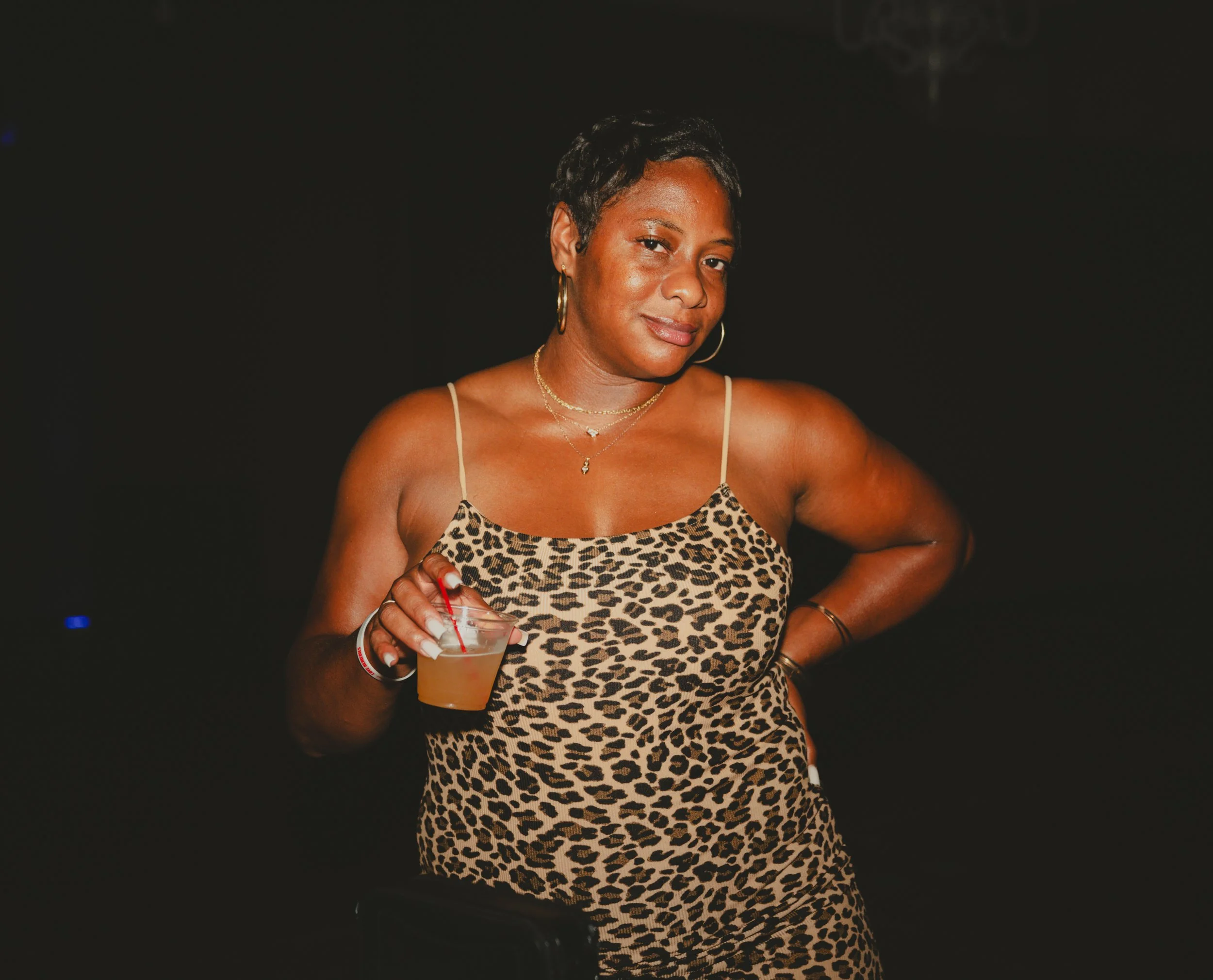 A woman with dark hair styled in twists, wearing hoop earrings, layered necklaces, and a leopard print spaghetti strap dress, holding a drink with a red straw, standing against a dark background.