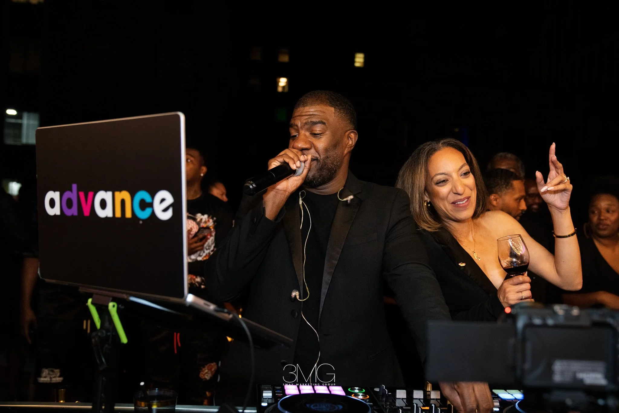 A man singing into a microphone at a party with a woman holding a glass of wine, surrounded by people and DJ equipment with an 'advance' logo.