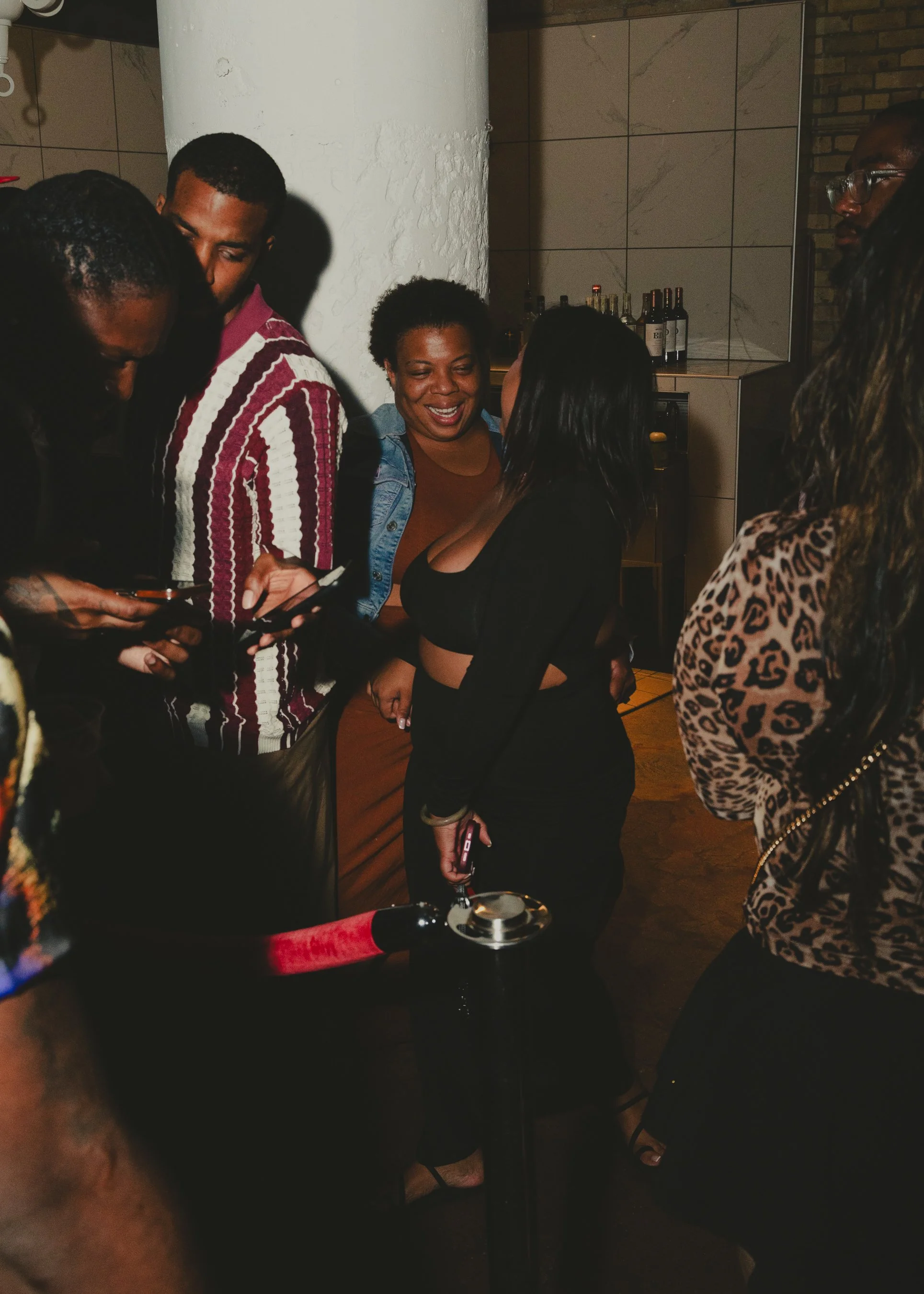 People at a social gathering, one person is holding a smartphone, others are talking and smiling. There is a bottle of champagne on the table.