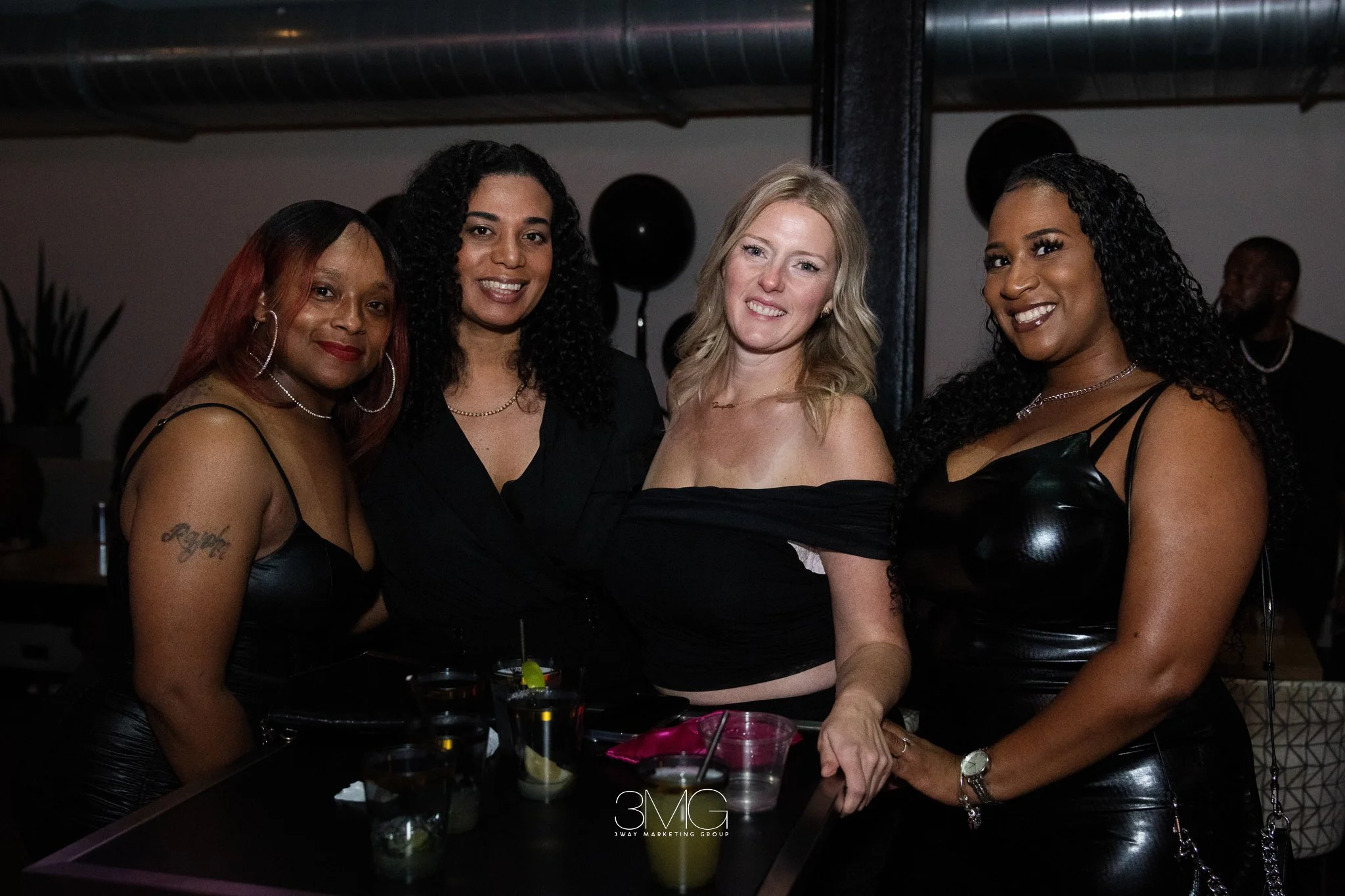 Four women smiling at a party or celebration, dressed in black, with drinks on the table in front of them, in a dimly lit indoor setting with black balloons and dark decor.